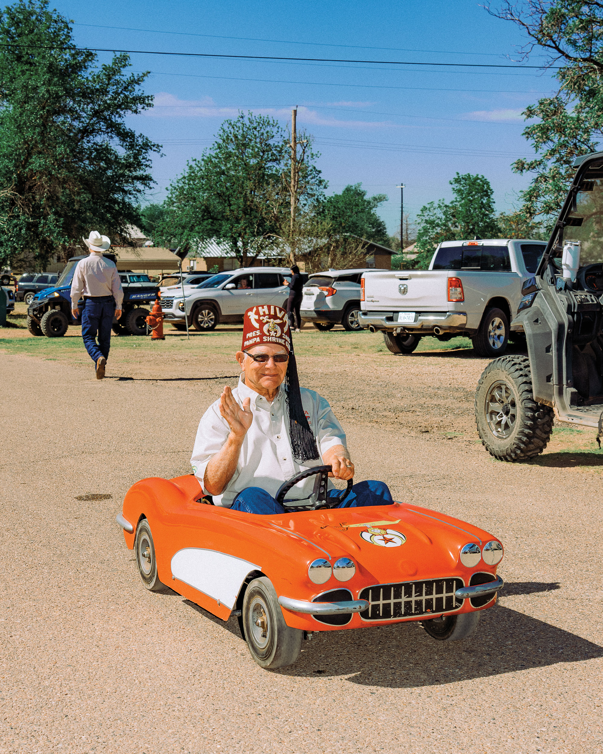 A man wearing a red hat with a tassel waves while driving a tiny red car