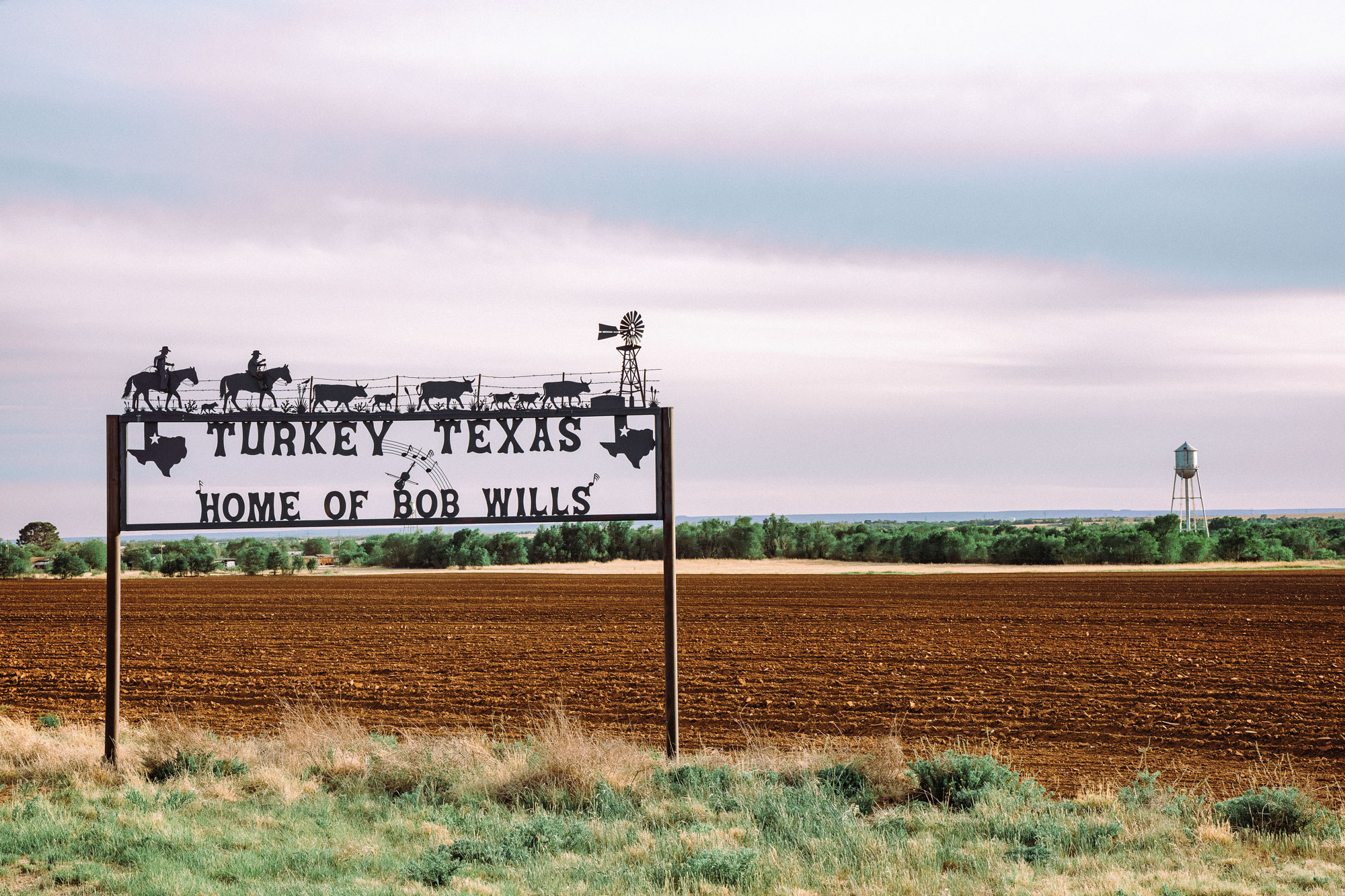 An iron sign that reads "Tukey, Texas Home of Bob Wills" stands in a field under an open sky