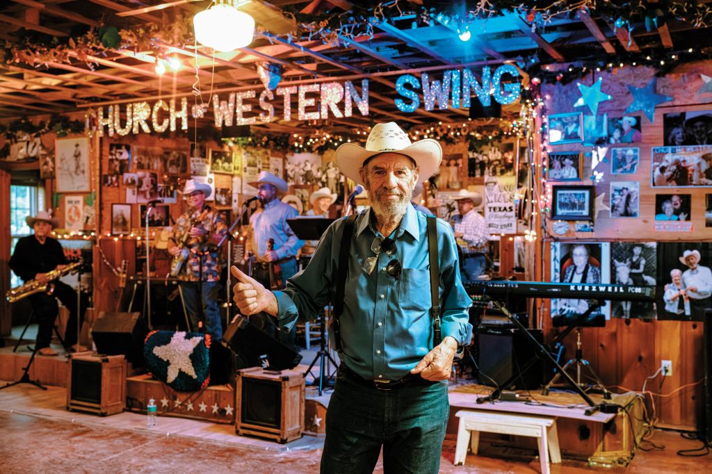 A man in a blue button down shirt and a cowboy hat holds a thumbs up inside a dance hall with a sign in the background that reads "Church of Western Swing"