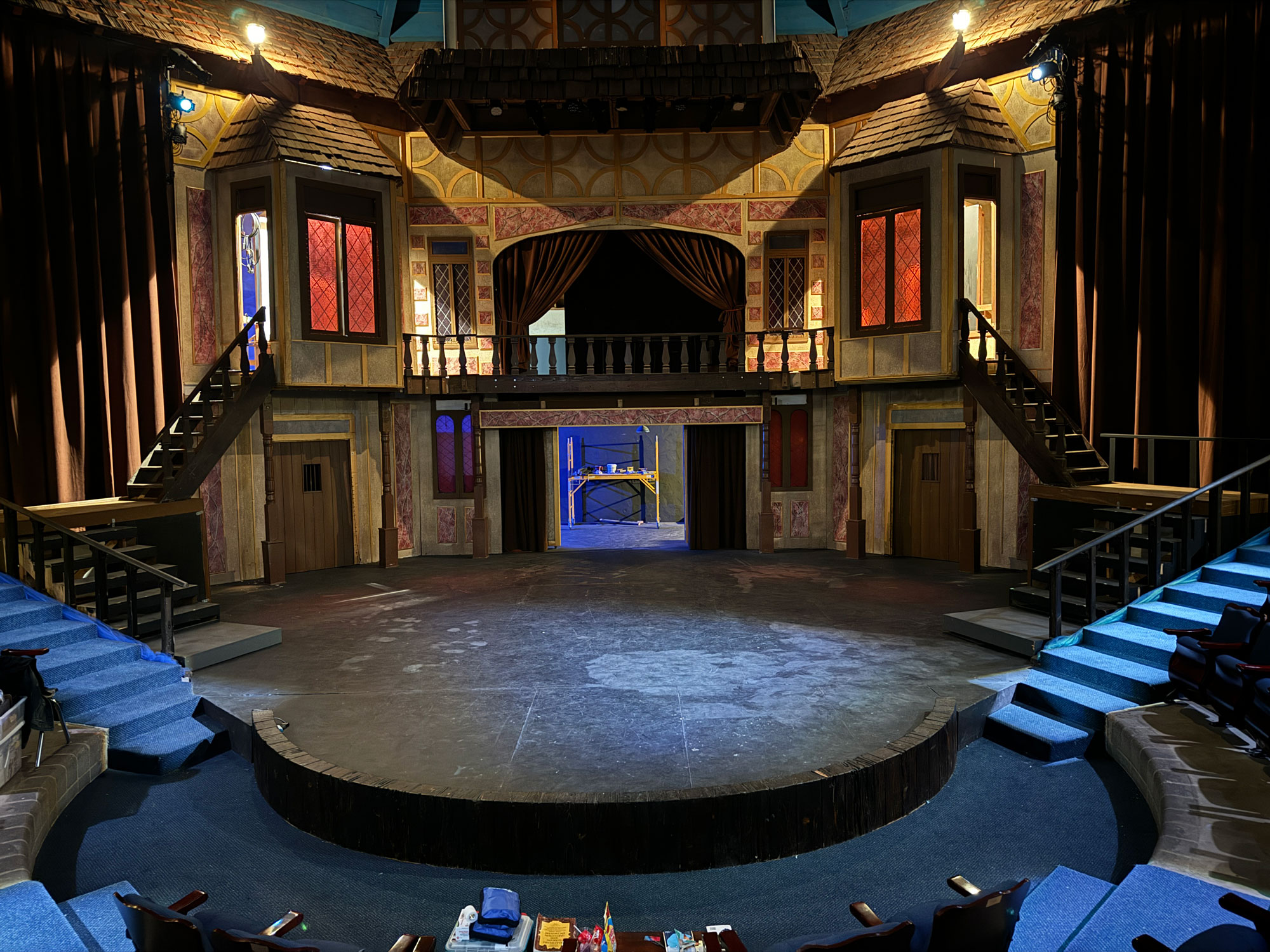 A circular stage in a theater with a set that has a second level with a balcony and red tinted windows.