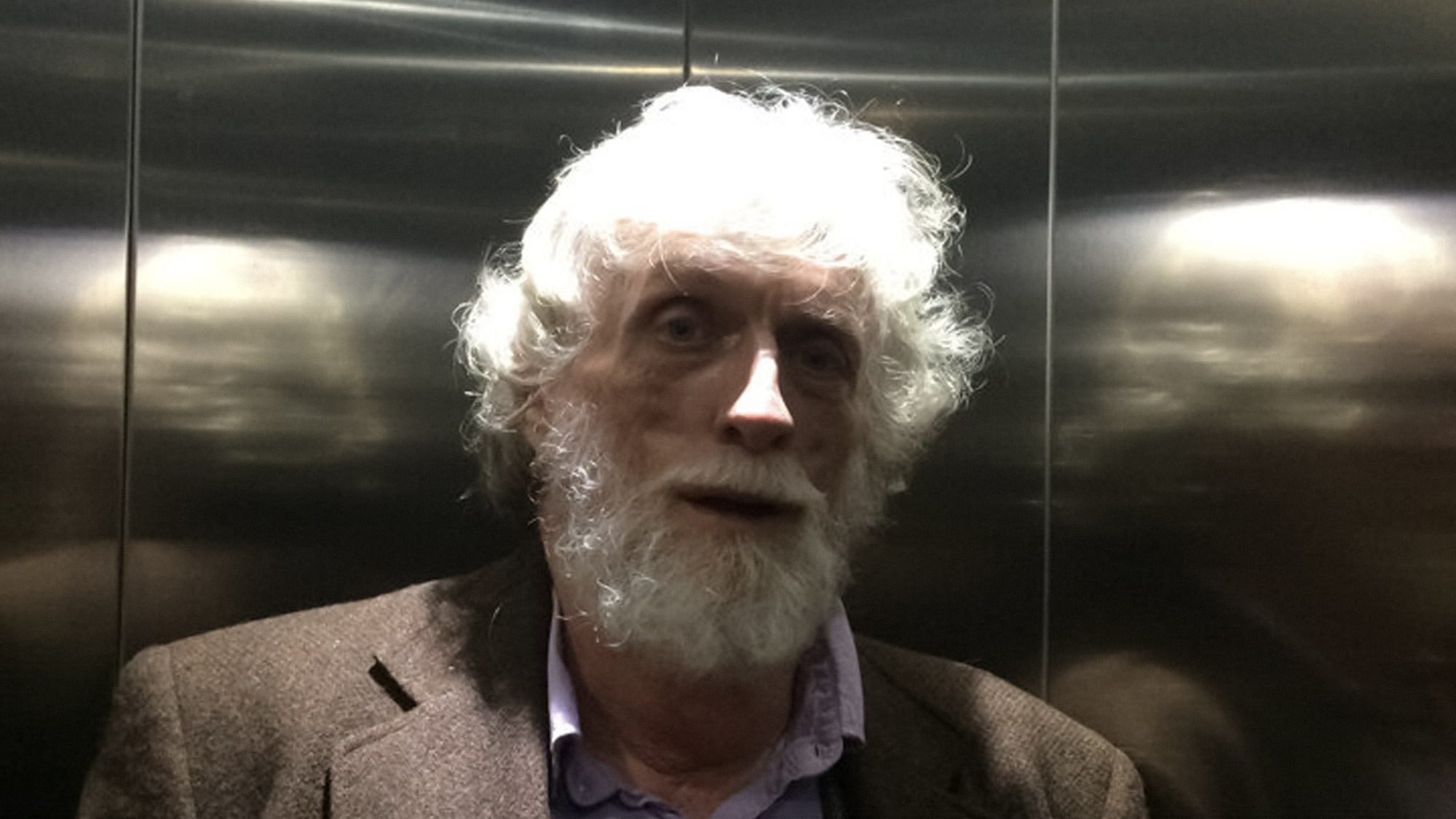 The artist James Magee, white-haired, looks directly at the camera as he stands in the corner of a reflective elevator.