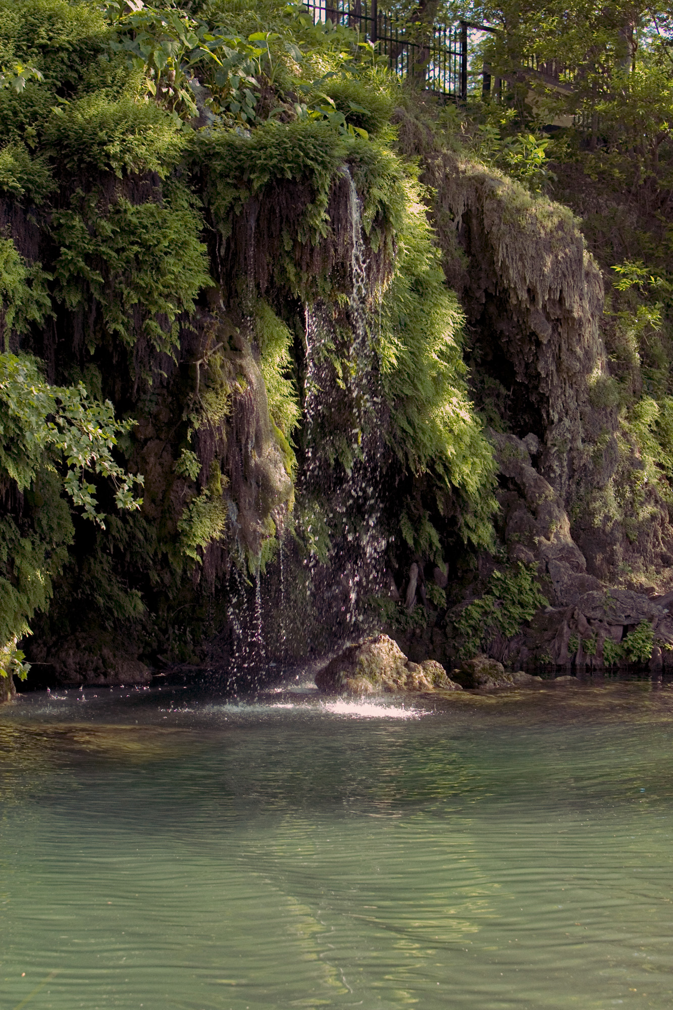 A waterfall drops water into a swimming hole near a rocky cliff and trees with green leaves