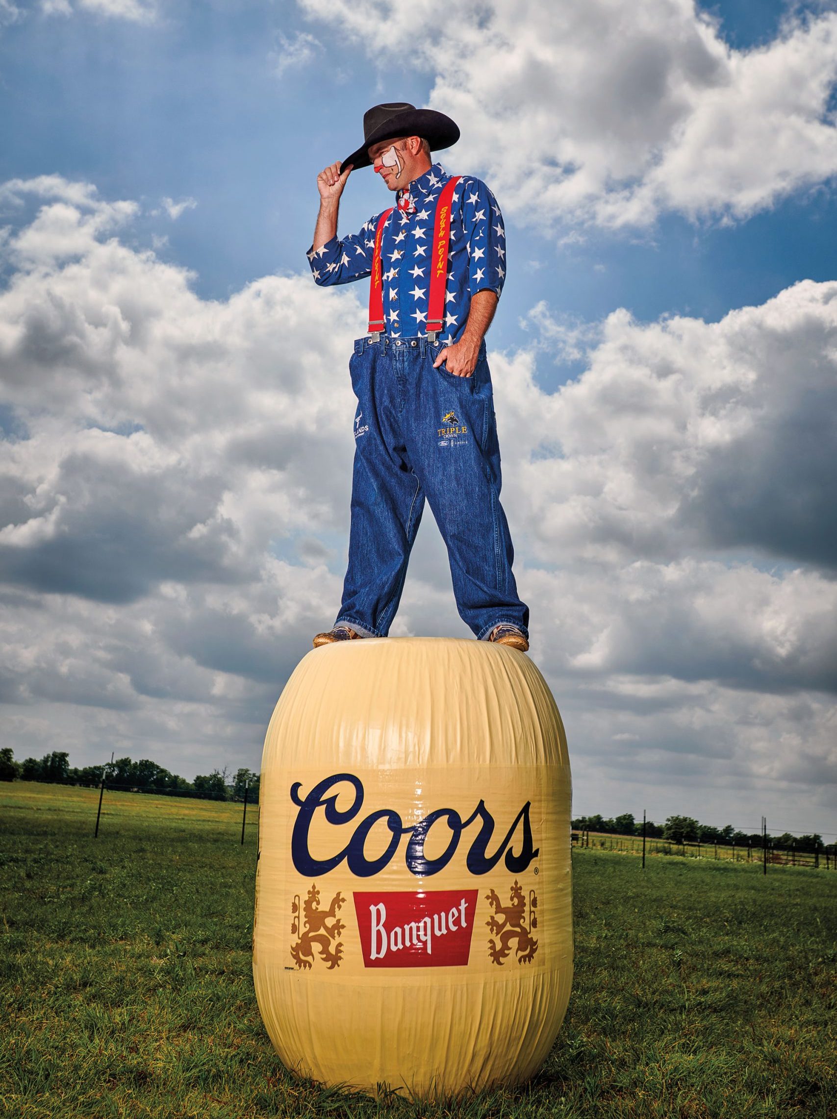 A man dressed as a rodeo clown in a large pair of denim jeans with red suspenders, a blue shirt with white stars, and a cowboy hat stands on a Coors Banquet barrel in a field.