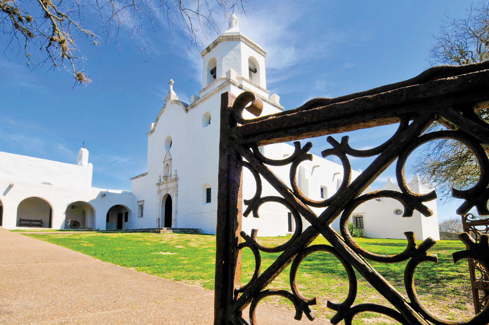 Mission Espiritu Santo, a white building with a cross over its door, sits on a green lawn with a black, rusted ornate fence in the foreground.