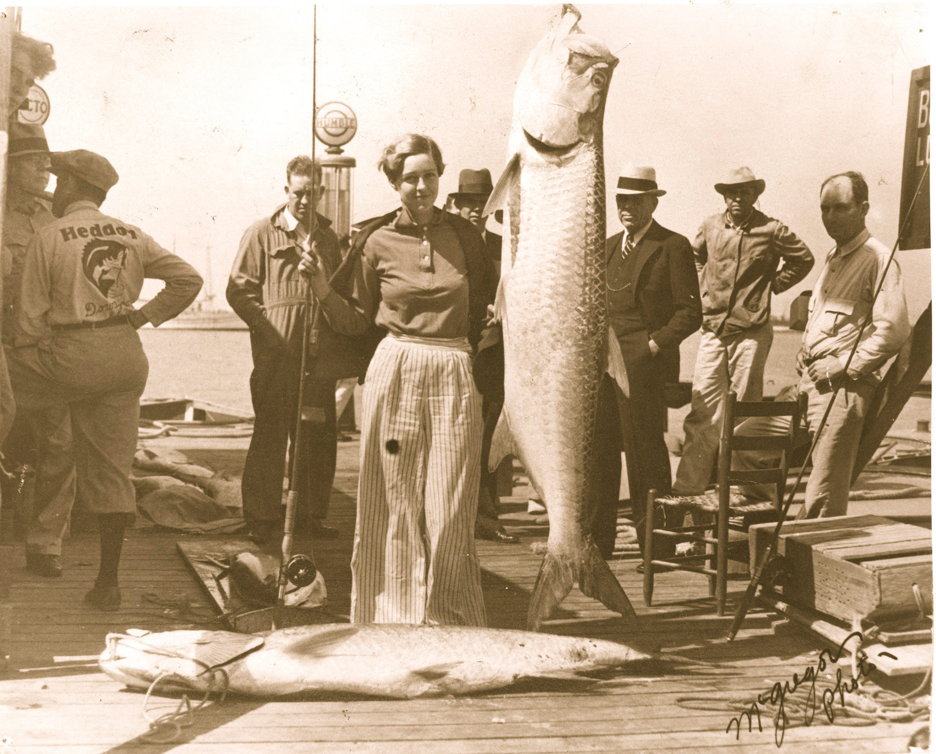 Black and white photo of Totsy Millican, posing in center next to massive fish hanging by a hook in its mouth, with men standing around her and her catch.