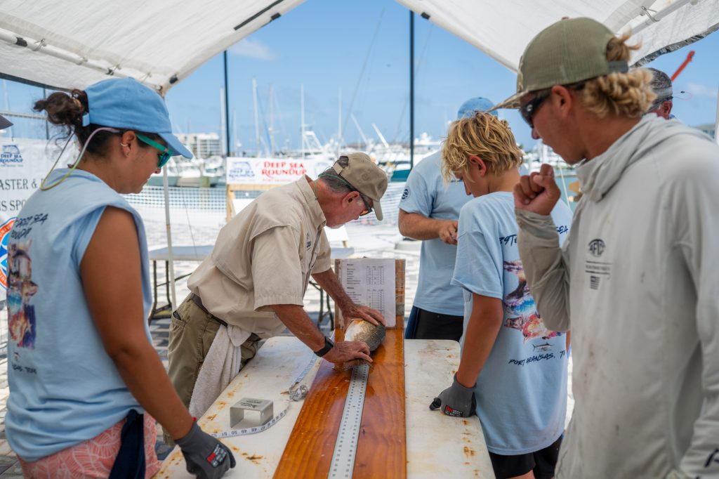 A man measures a fish placed atop a long ruler on a tabletop as a few people in the same T-shirts and a man in a gray hoodie stand around him and watch.