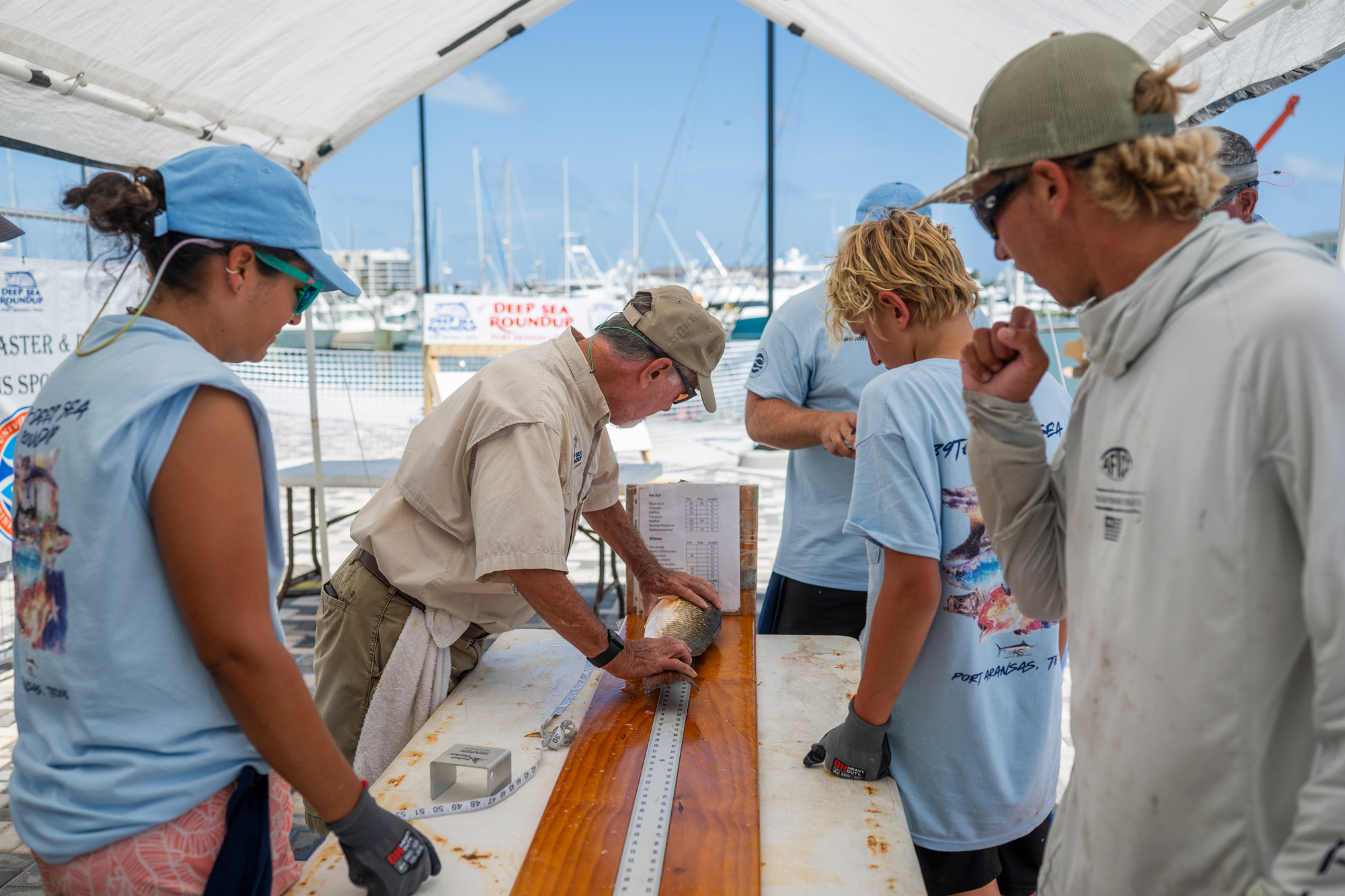 A man measures a fish placed atop a long ruler on a tabletop as a few people in the same T-shirts and a man in a gray hoodie stand around him and watch.