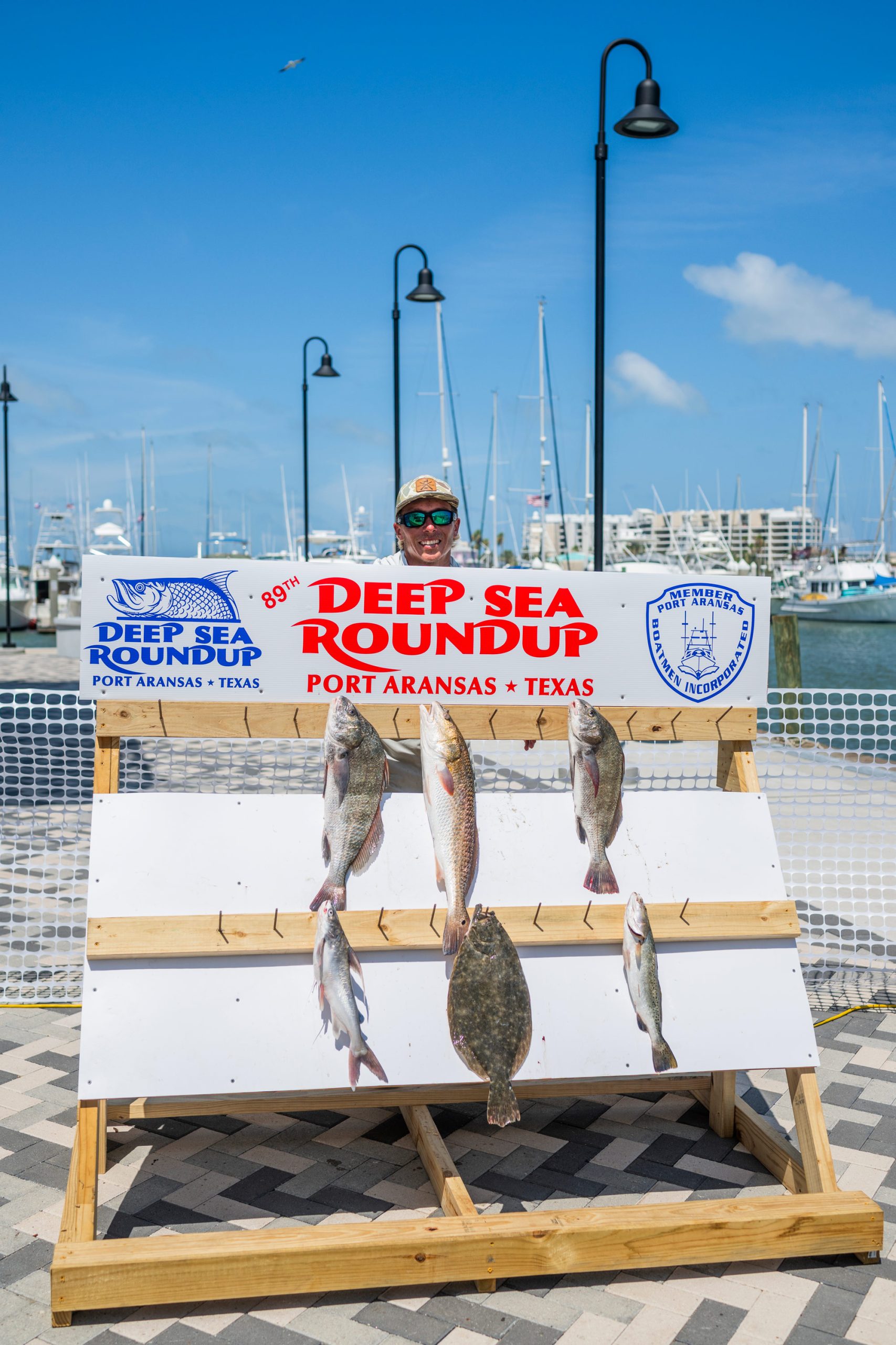 A man stands behind a large wooden stand with the words Deep Sea Roundup on a sign and six fish affixed to it