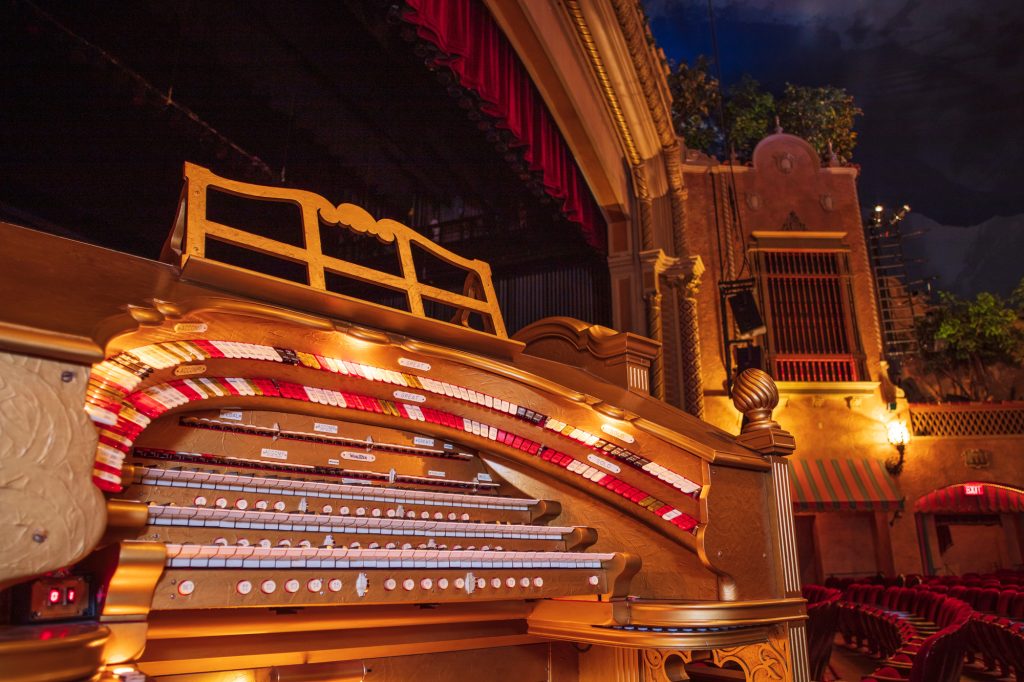 A large gold pipe organ with three rows of keys and numerous buttons inside a theater with red seats and a red curtain