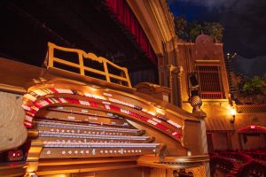 A large gold pipe organ with three rows of keys and numerous buttons inside a theater with red seats and a red curtain