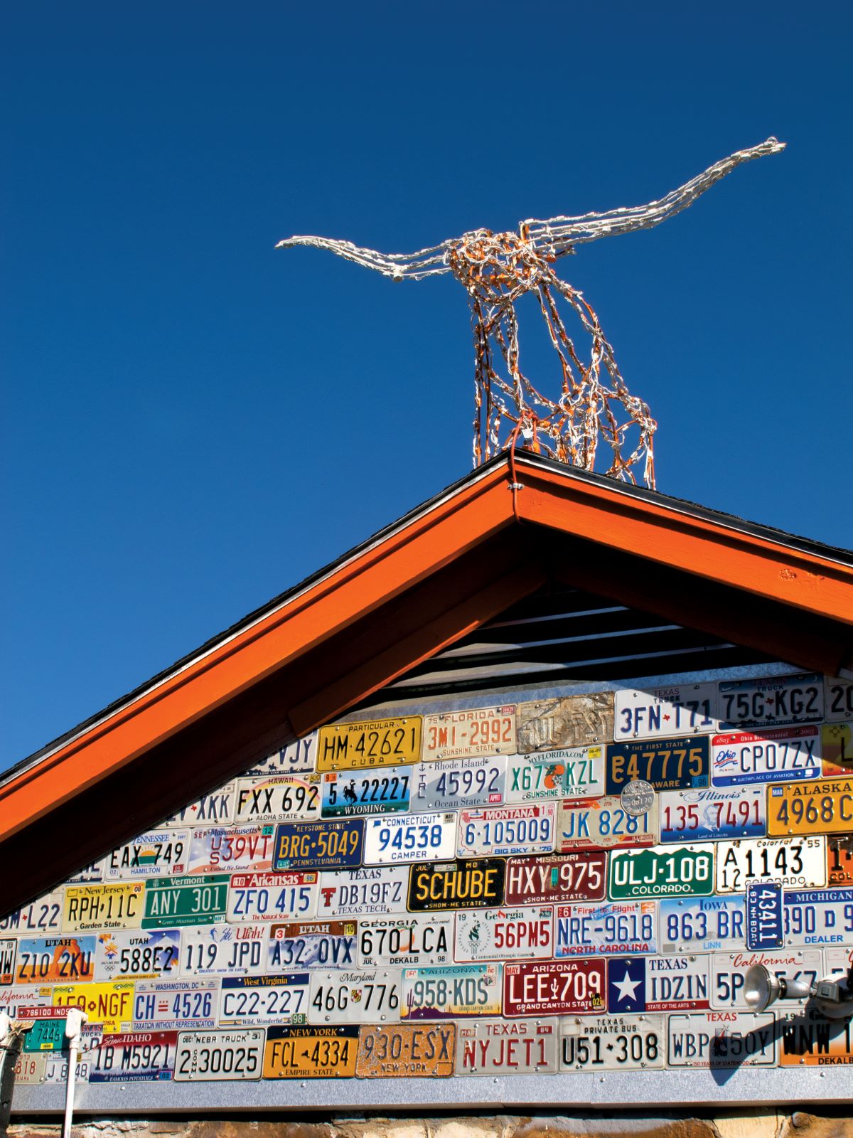 Wire longhorn perched on the peak of the roof of the Little Longhorn Saloon above a collage of different colored license plates on the exterior of the building