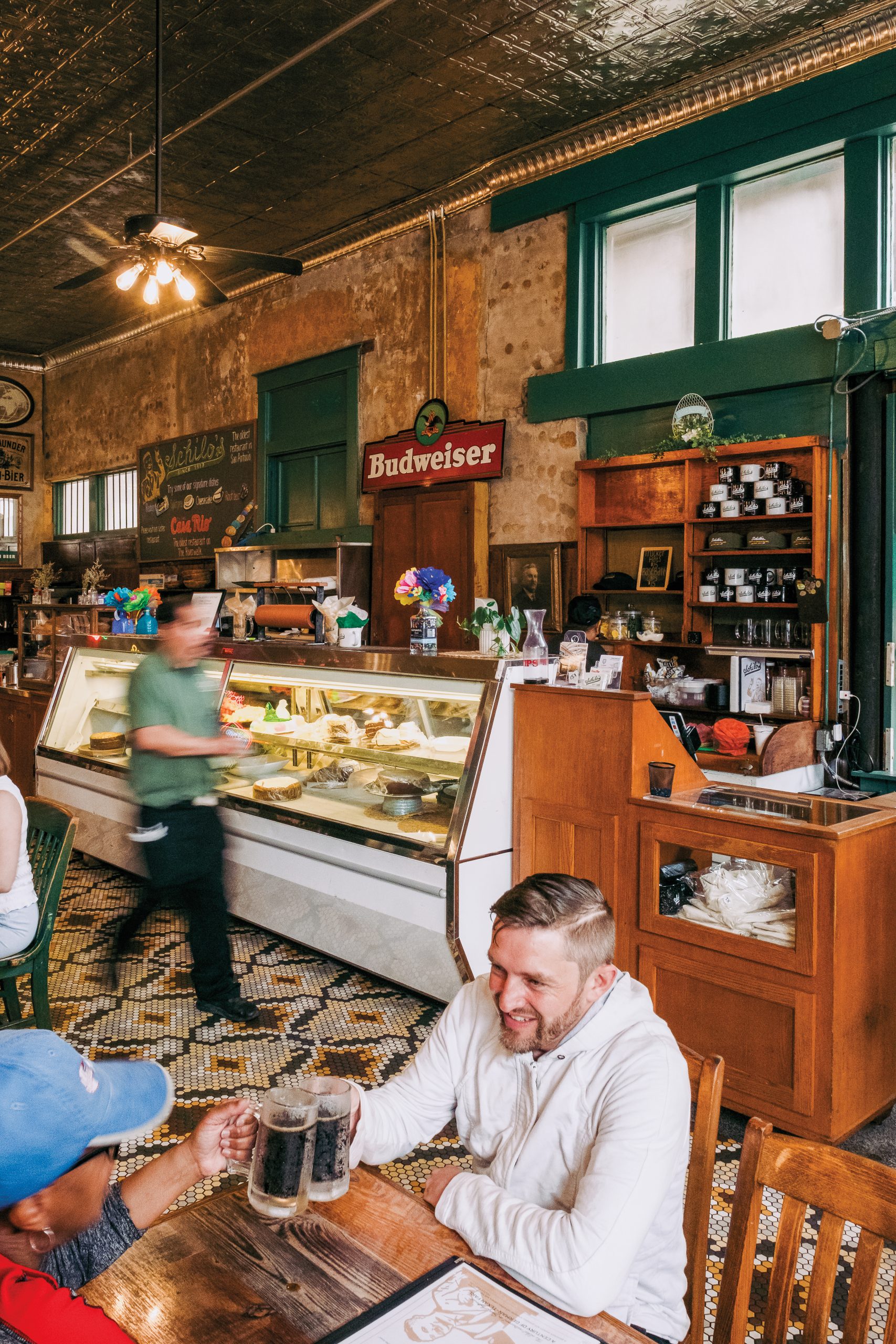 People cheers with mugs of root beer inside a green and brown shop with a pastry case in the background