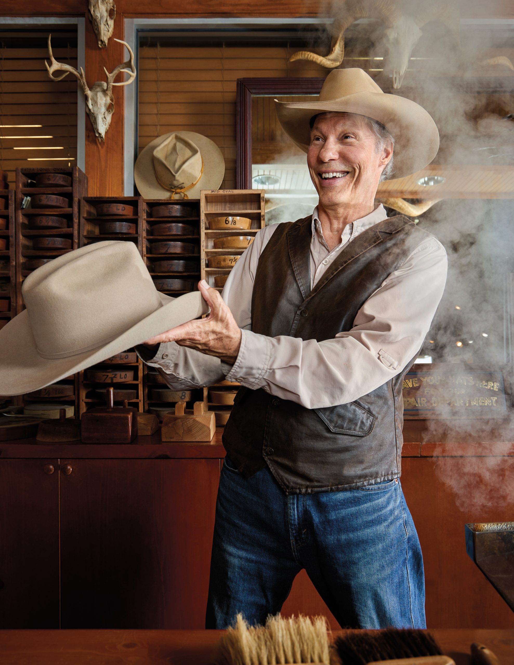 A man in a cowboy hat, vest, and jeans holds an ivory cowboy hat while smiling and standing in front of a cloud of steam.