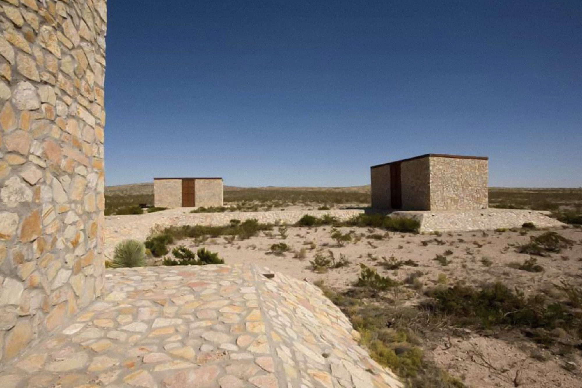 A partial view of three buildings of James Magee's The Hill made of rough-cut rock, which stand on desert grassland.