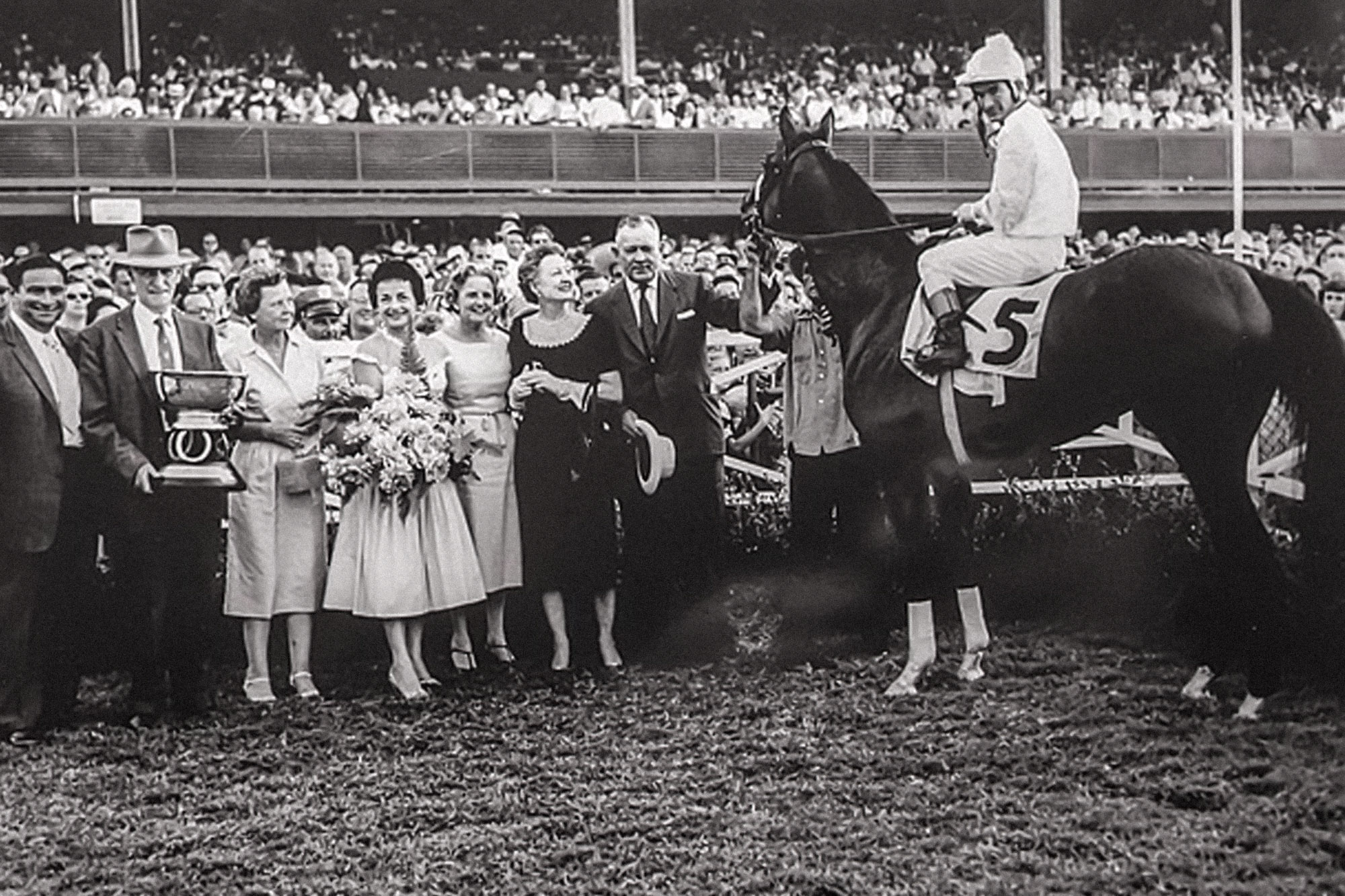 A black & white photograph of a jockey aboard a horse poses victorious in front of a large crowd.