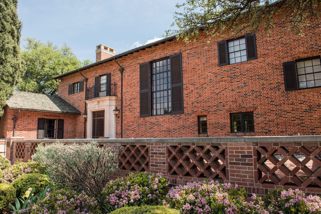 A large red-brick estate during the daytime, fronted with vibrant foliage and bushes.