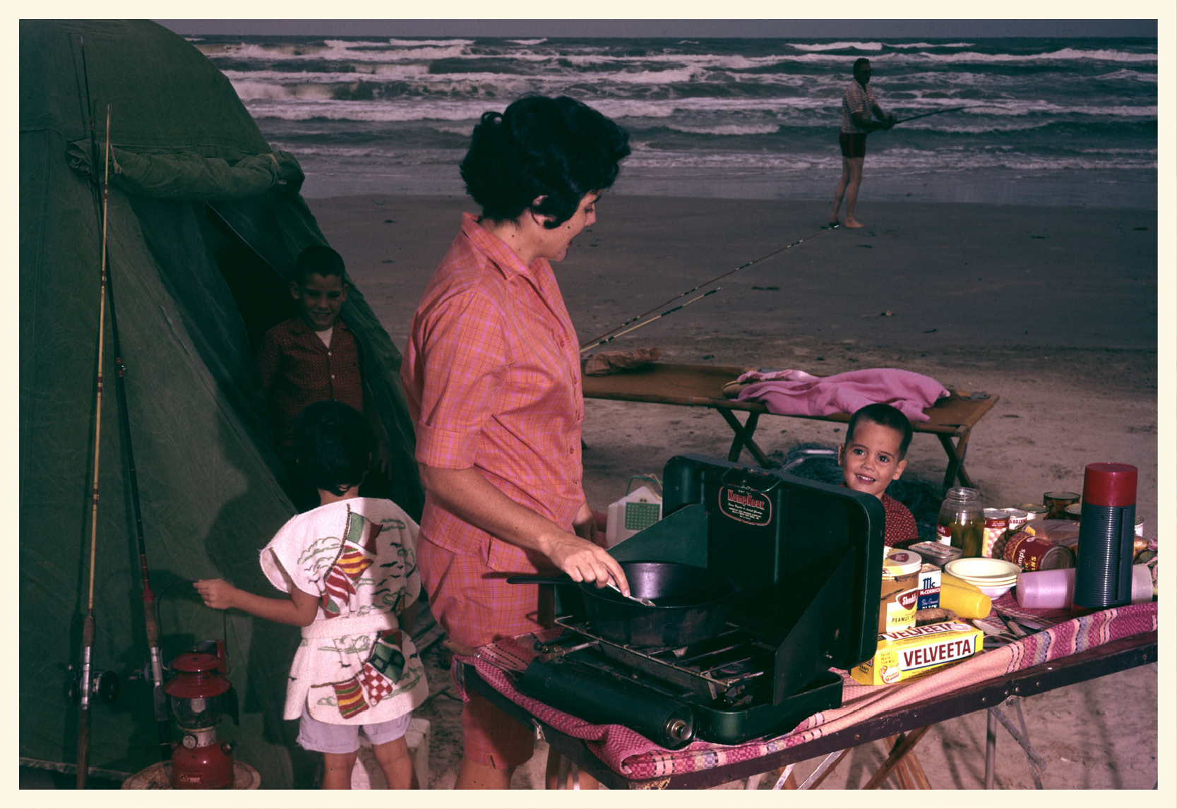 A vintage photo of a woman in a pink outfit cooking on a grill on the beach surrounded by children and a tent.
