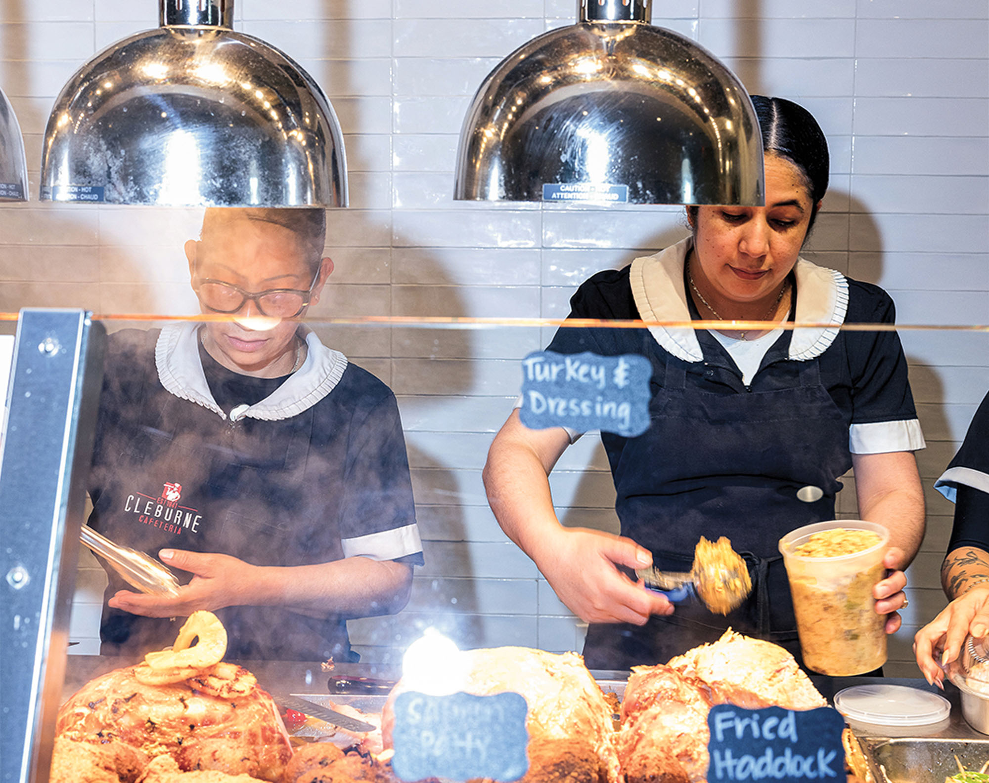 Two women dressed in black uniforms working behind a glass-fronted cafeteria line serving food and preparing containers.