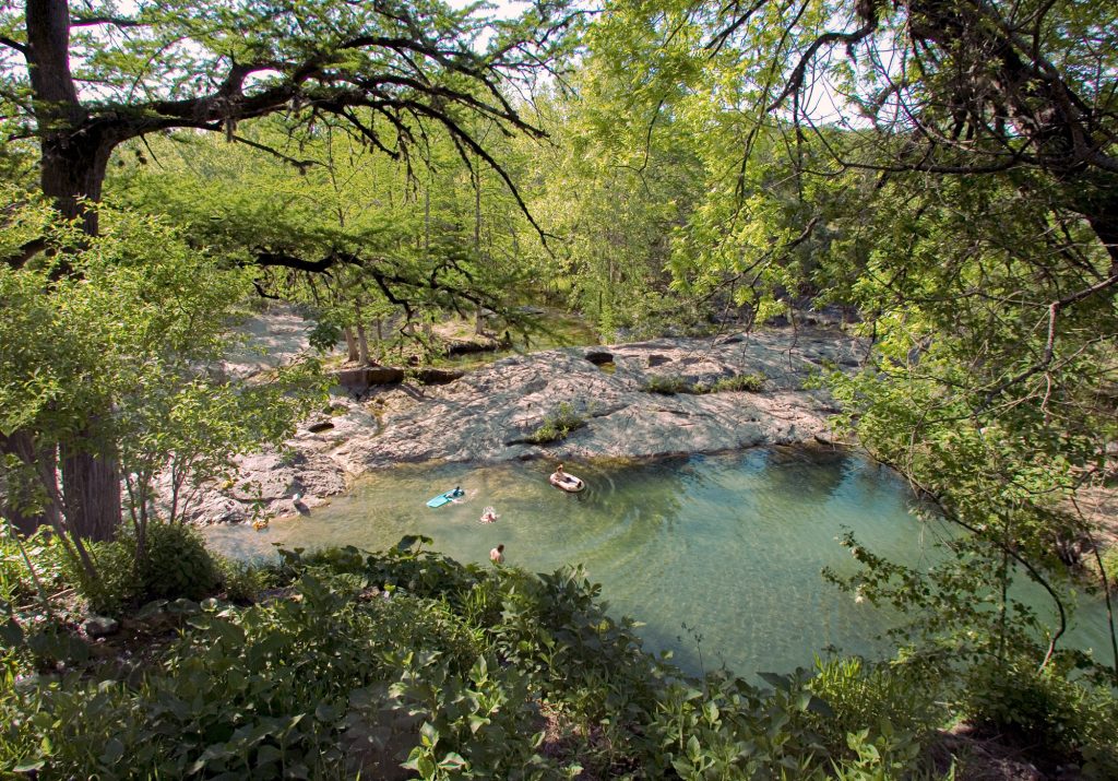 A swimming hole with four people in the water, surrounded by rocks and trees with bright green leaves
