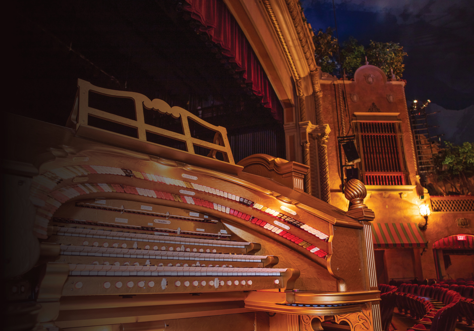 A large gold pipe organ with three rows of keys and numerous buttons inside a theater with red seats and a red curtain