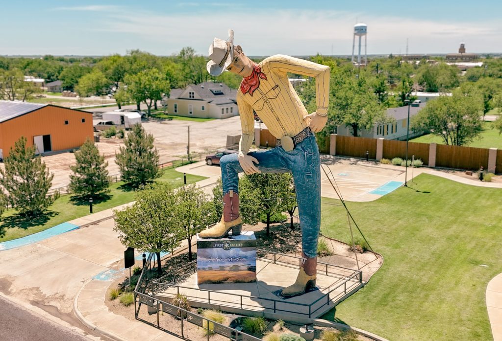 Tex Randall, a giant cowboy statue, has his hand on its hip and boot propped up on a sign that reads The Spirit of West Texas in a park.
