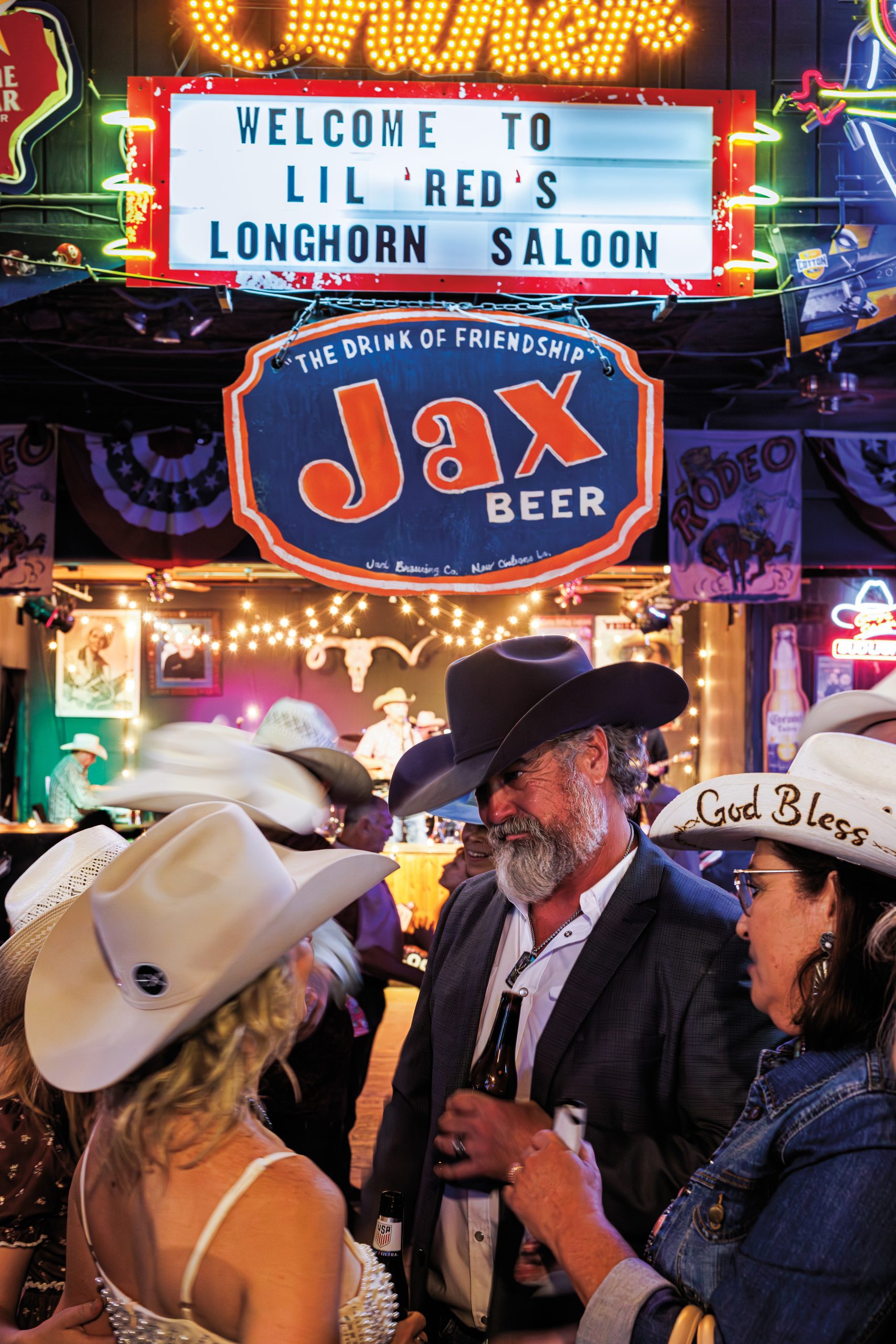A man in a black cowboy hat with a thick grey beard stands in a group with three women in ivory cowboy hats, one of which reads "God Bless" on the side. They have beers in their hands. Twinkle lights and a neon marquee is in the background. The sign reads "Welcome to Lil Red's Longhorn Saloon."