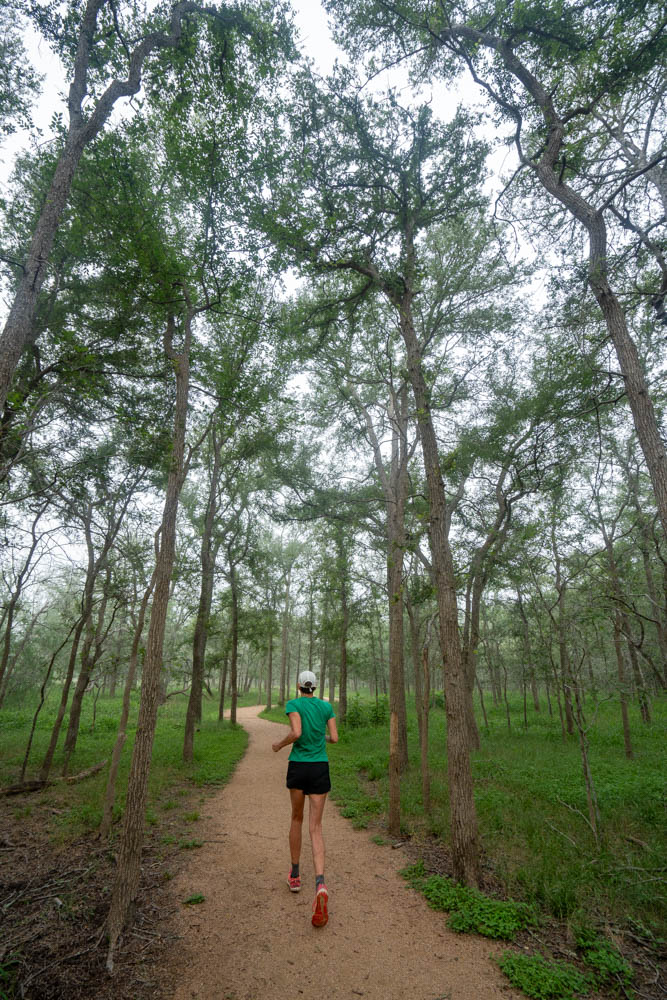 A person in a green shirt and black shorts running on a trail through a wooded area
