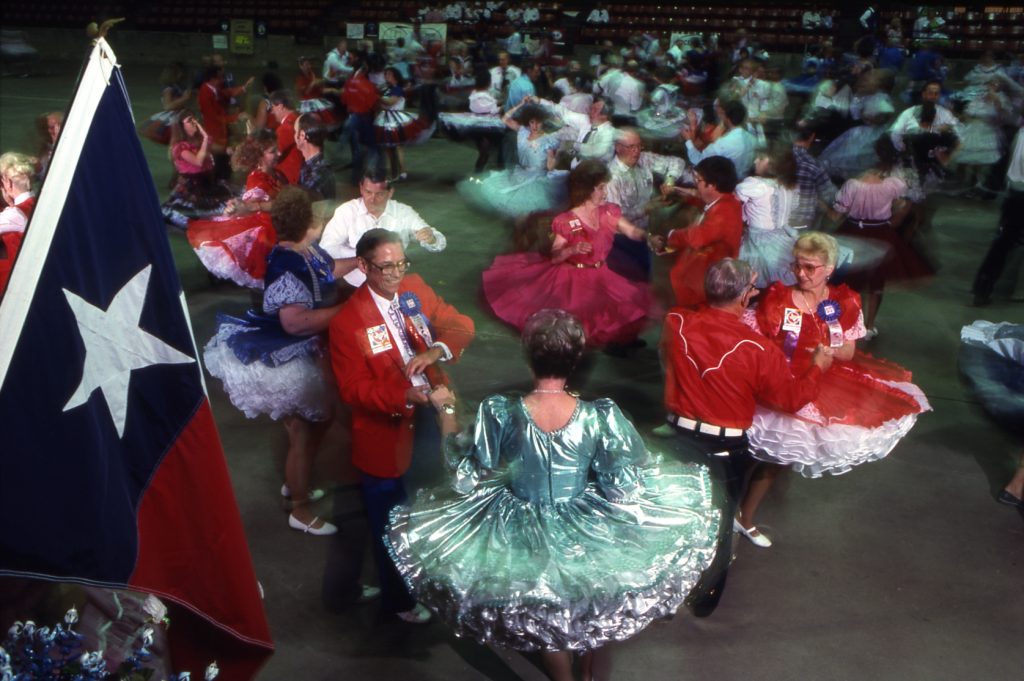 People in red and blue outfits dance beneath a large Texas flag in a large dance hall