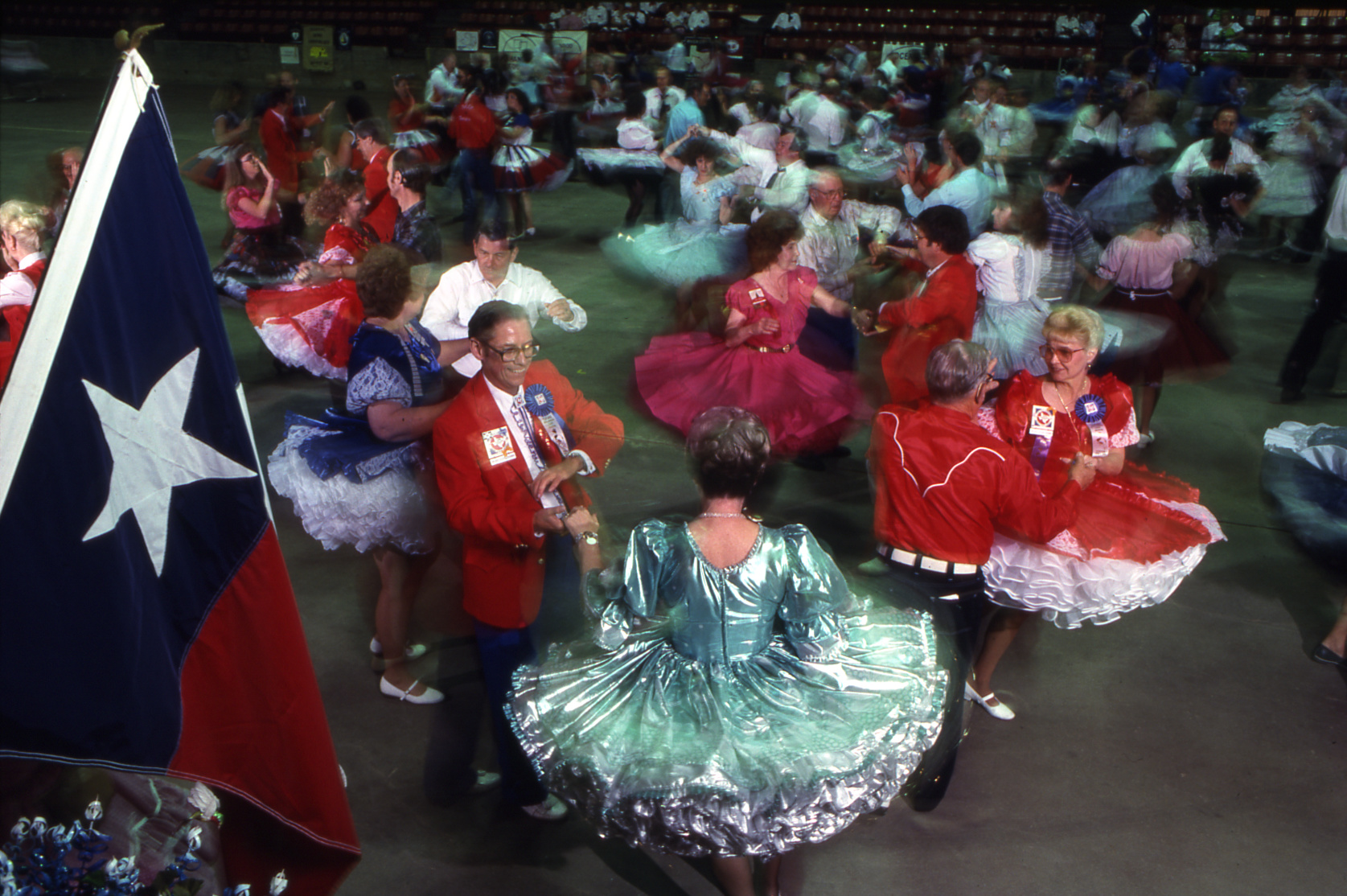 People in red and blue outfits dance beneath a large Texas flag in a large dance hall