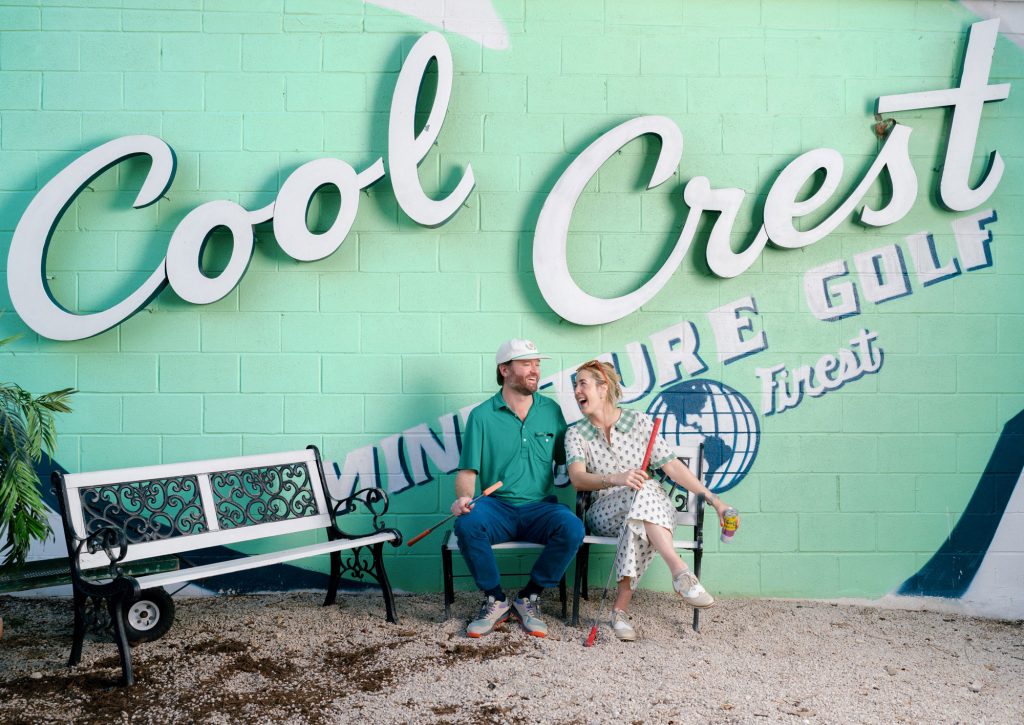 A man in a green shirt and white baseball cap sits next to a laughing woman in a white dress in front of a giant Cool Crest Miniature Golf sign painted on a wall.