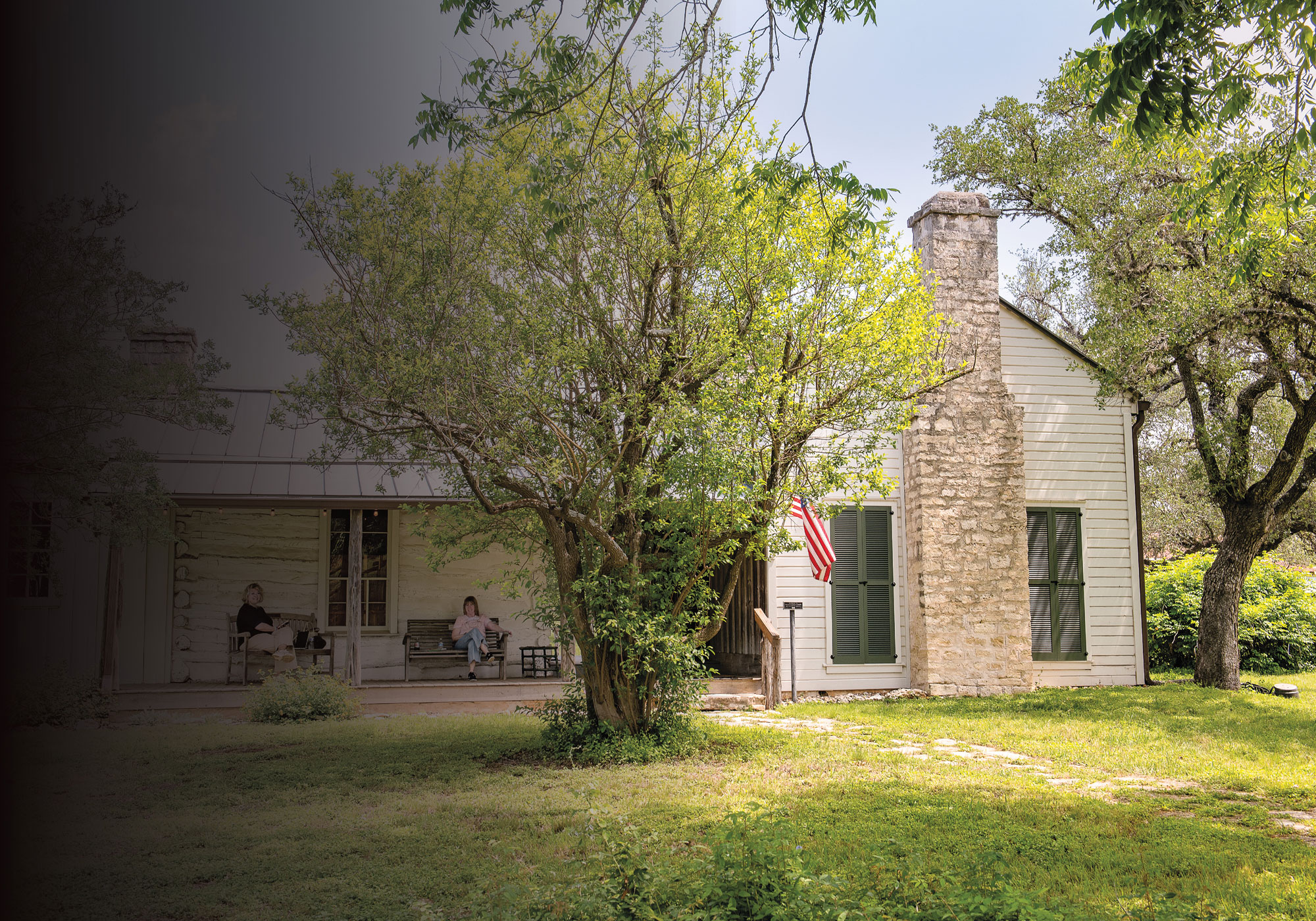 Two women sit on the porch of a large white home in a modernized rustic ranch style. Its grassy front yard features multiple medium-sized trees.