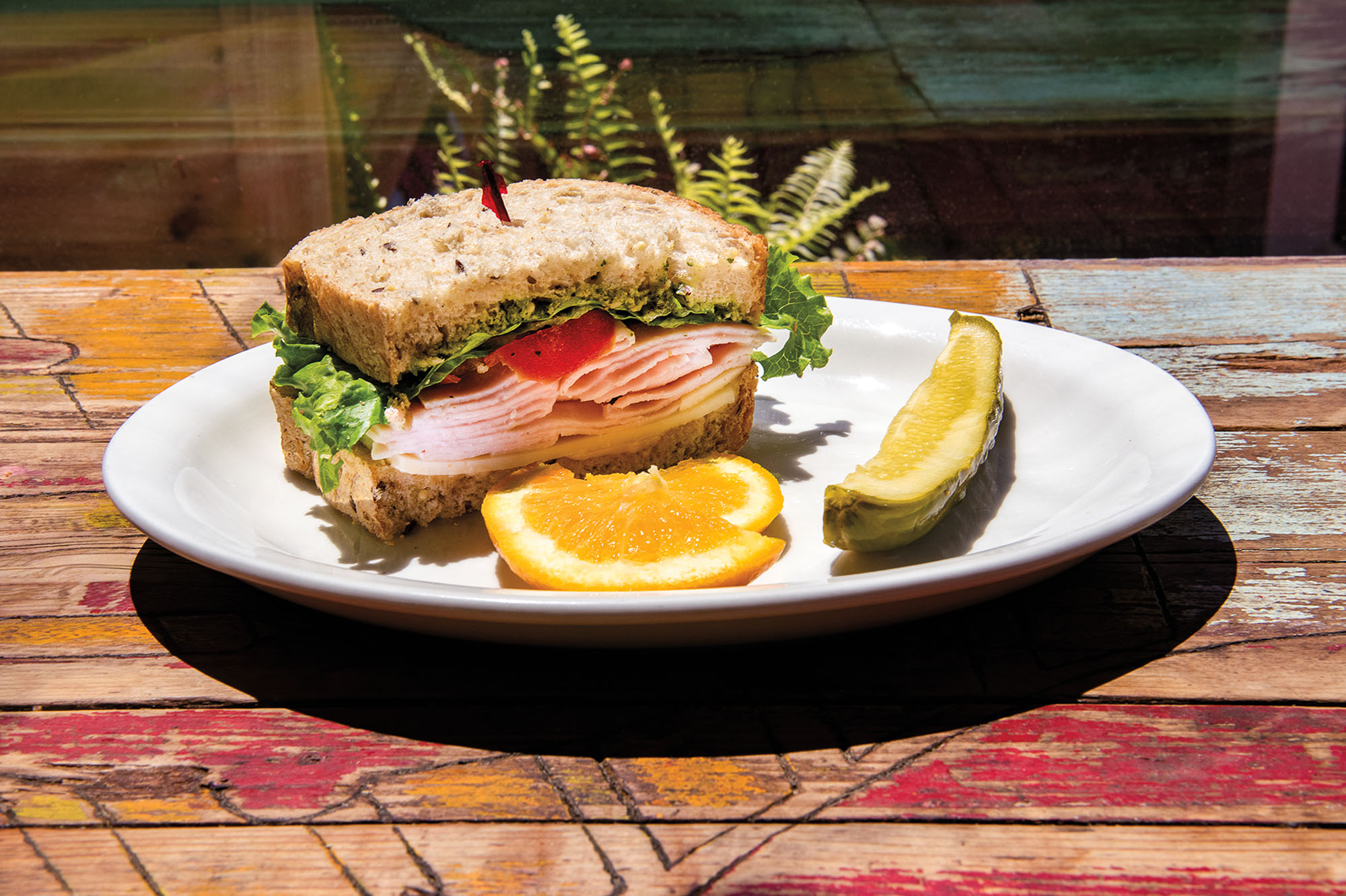 A club sandwich, pickle, and wedge of orange plated on a white dish on a rustic table.