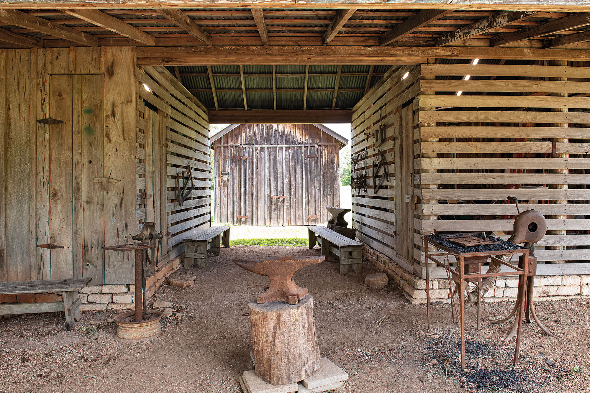 A rustic outdoor smithy made of brown wooden panels featuring a central anvil on a dirt floor.