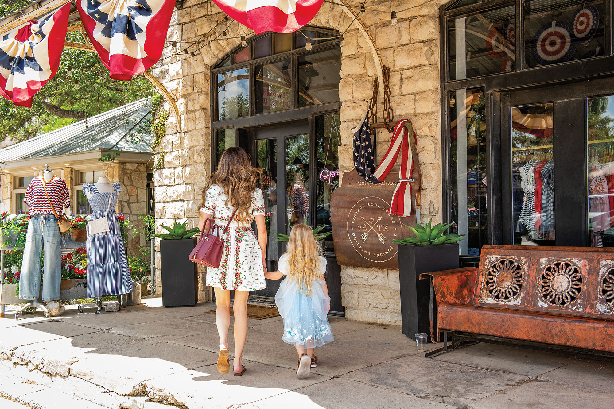 A woman and young girl holding hands as they walk down a cozy street of vintage storefronts during the daytime.