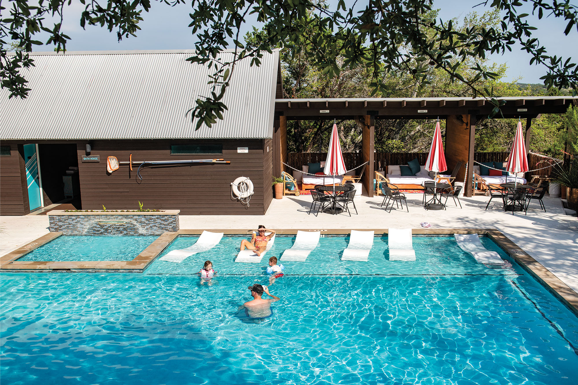 A young family of four enjoying their daytime in the large pool at Lucky Arrow Retreat in Dripping Springs. The pool features curved white lounge chairs and a hot tub, with brown buildings and red-white umbrellas seen in the background.