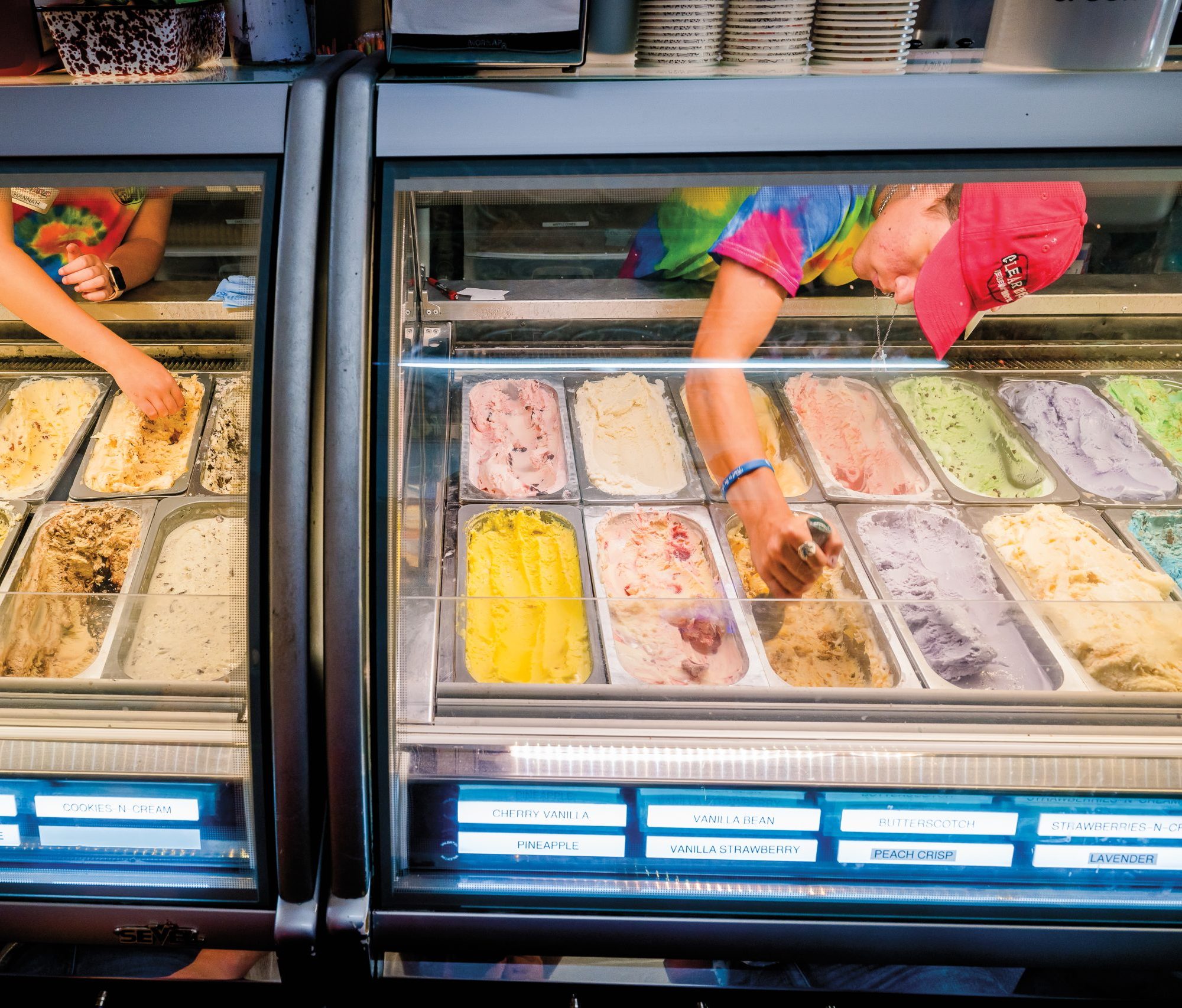 A young worker, wearing a pink hat, scooping a brown-colored ice cream from among other vibrant options, behind a glass counter.