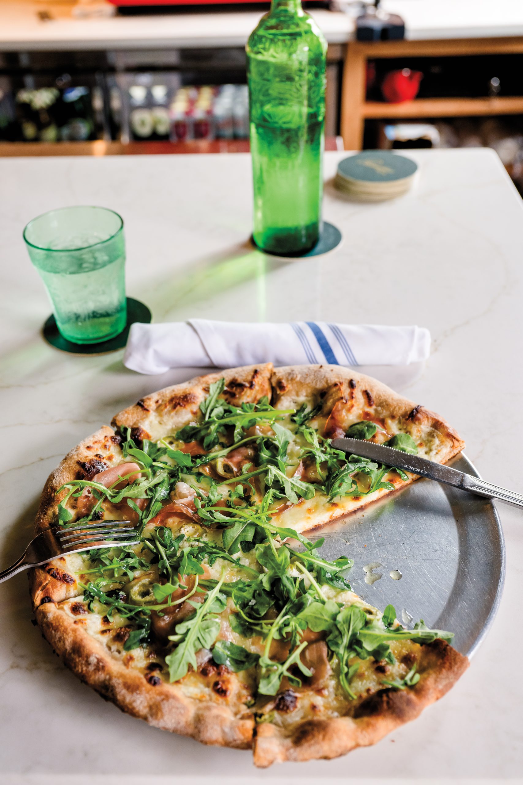 A partially-eaten pizza topped with arugula, prosciutto, and olives served on a metal plate with a fork and knife. A green glass bottle and green water glass appear behind.