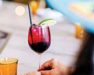 A magenta-colored drink served in a clear wine glass with a wedge of lime, with its base lightly gripped by a woman, on a marble table.