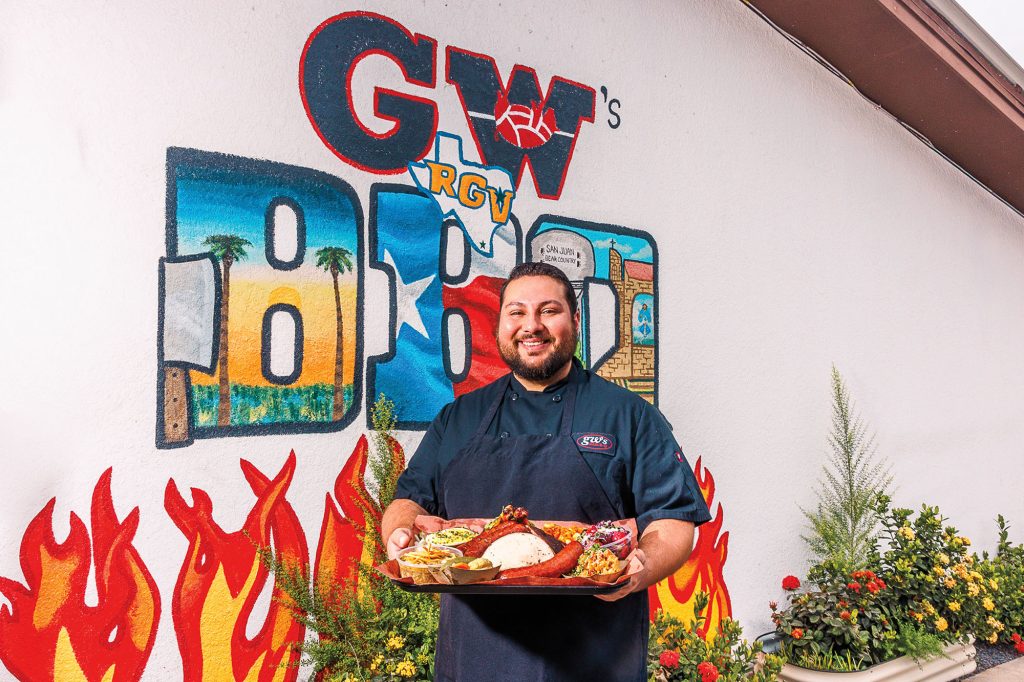 A smiling chef holds a plate containing a spread of his cooking, including barbecue meats and sides. Behind him, a white wall displays a painted "GW’s BBQ" logo. The mural also features flames at the bottom and a small Texas silhouette with "RGV" in front. Potted plants are displayed against the wall.