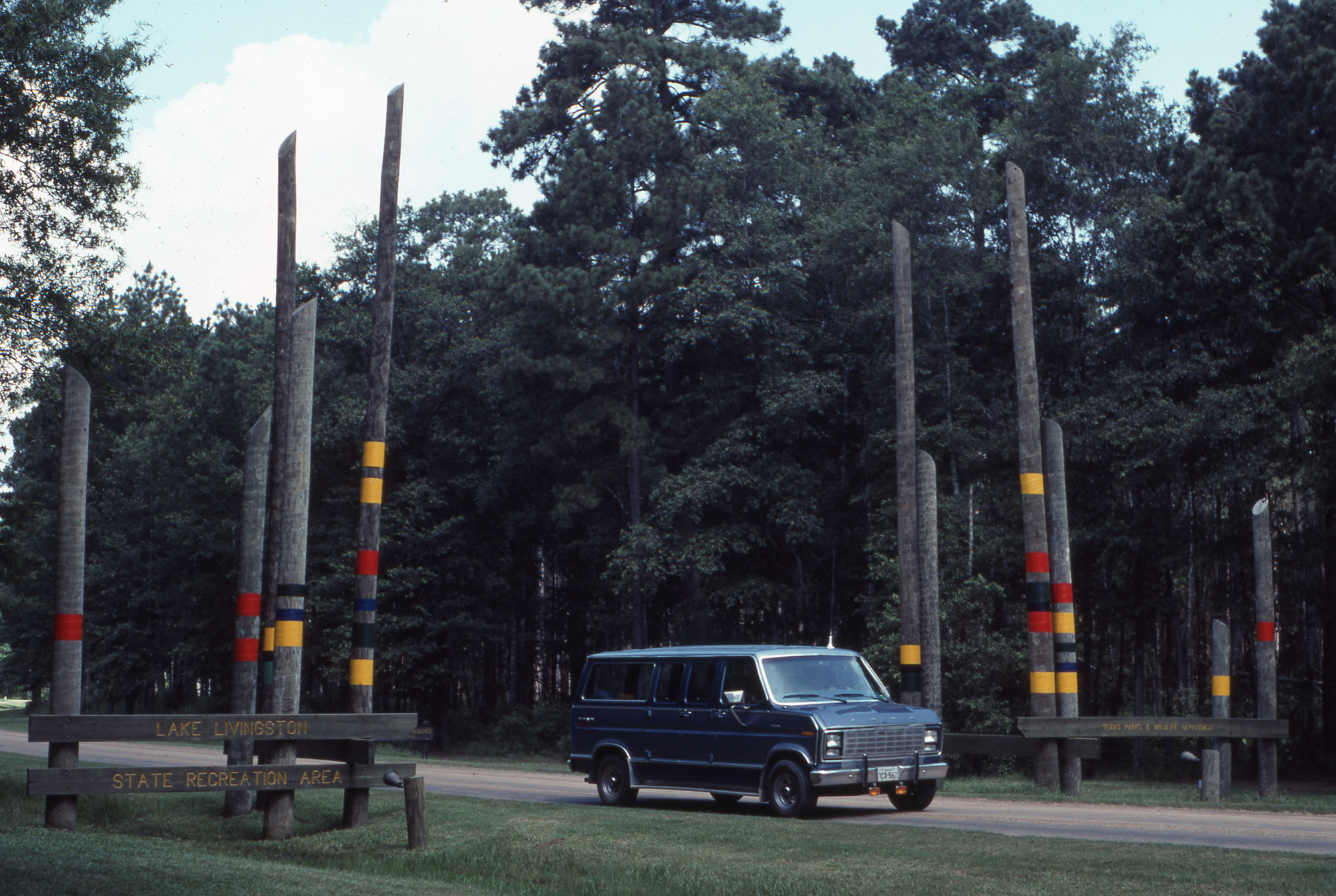 A blue minivan on a road through a forest with a wooden sign that reads Lake Livingston Recreation Area