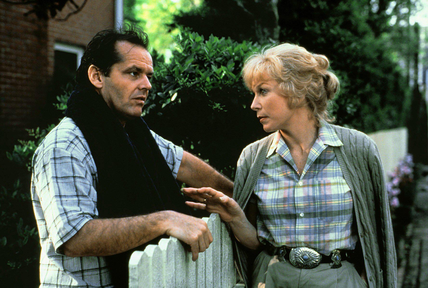 A man and a woman stand on either side of a white picket fence, both with pensive stares