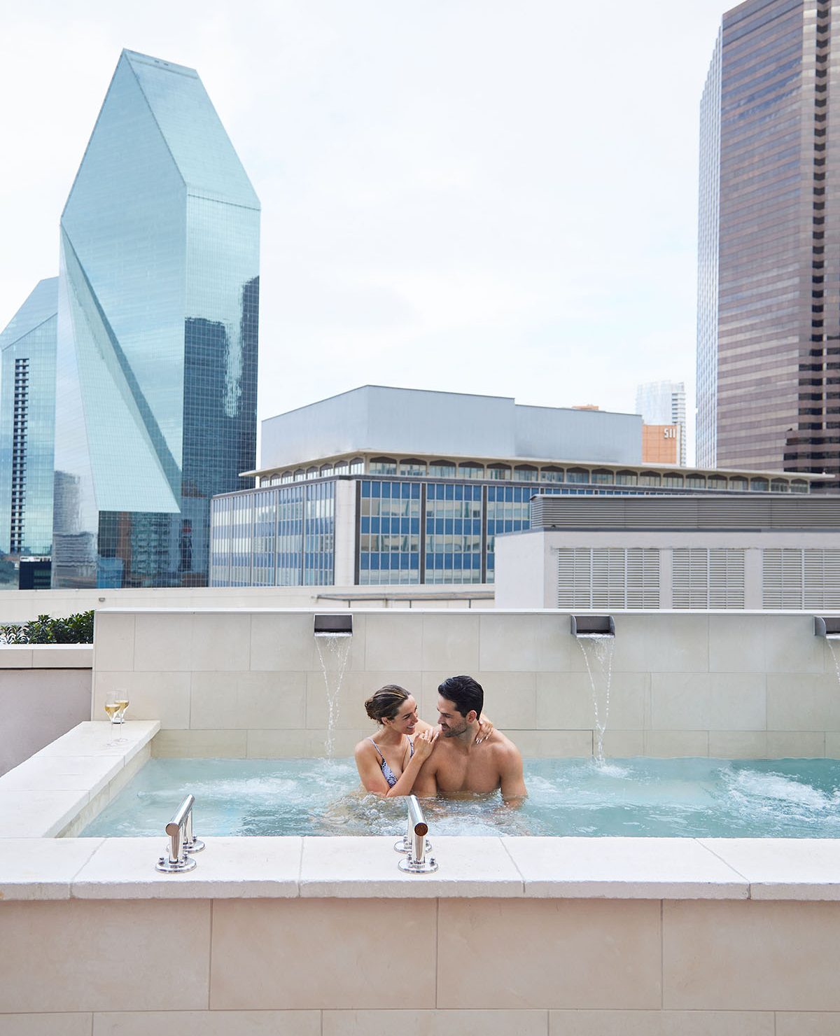 A couple steams in a hot tub overlooking a glass skyscraper.