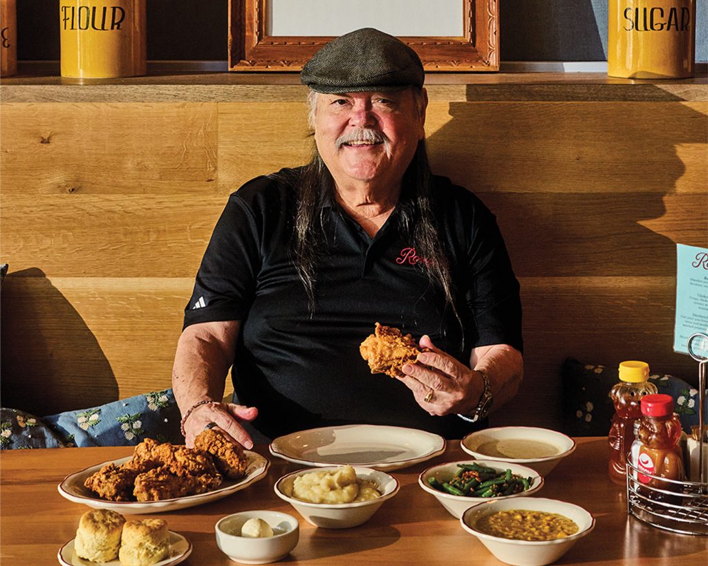 A smiling man wearing a black polo and flat cap holds a piece of fried chicken in a homestyle restaurant interior. A framed vintage portrait of a couple, as well as two yellow jars labeled "FLOUR" and "SUGAR," appear behind him. He sits at a table featuring a spread of Southern cooking.