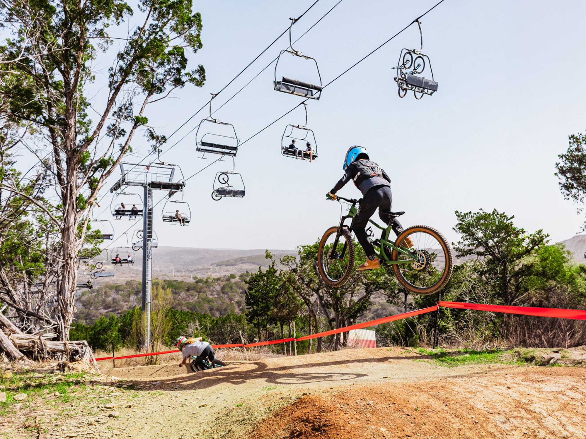 A mountain biker jumps a dirt hill with a chairlift above him.
