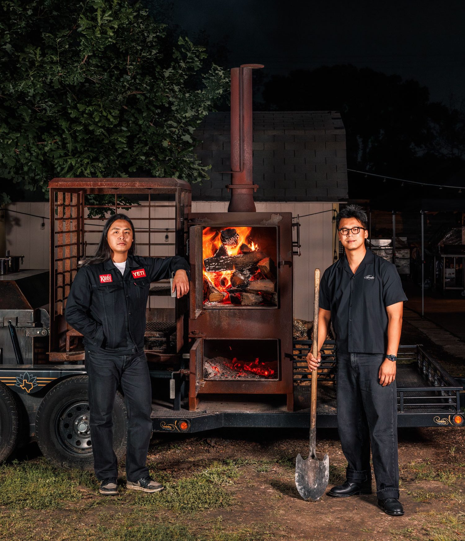 Two Asian men in black outfits stand on either side of a lit smoker outside at night