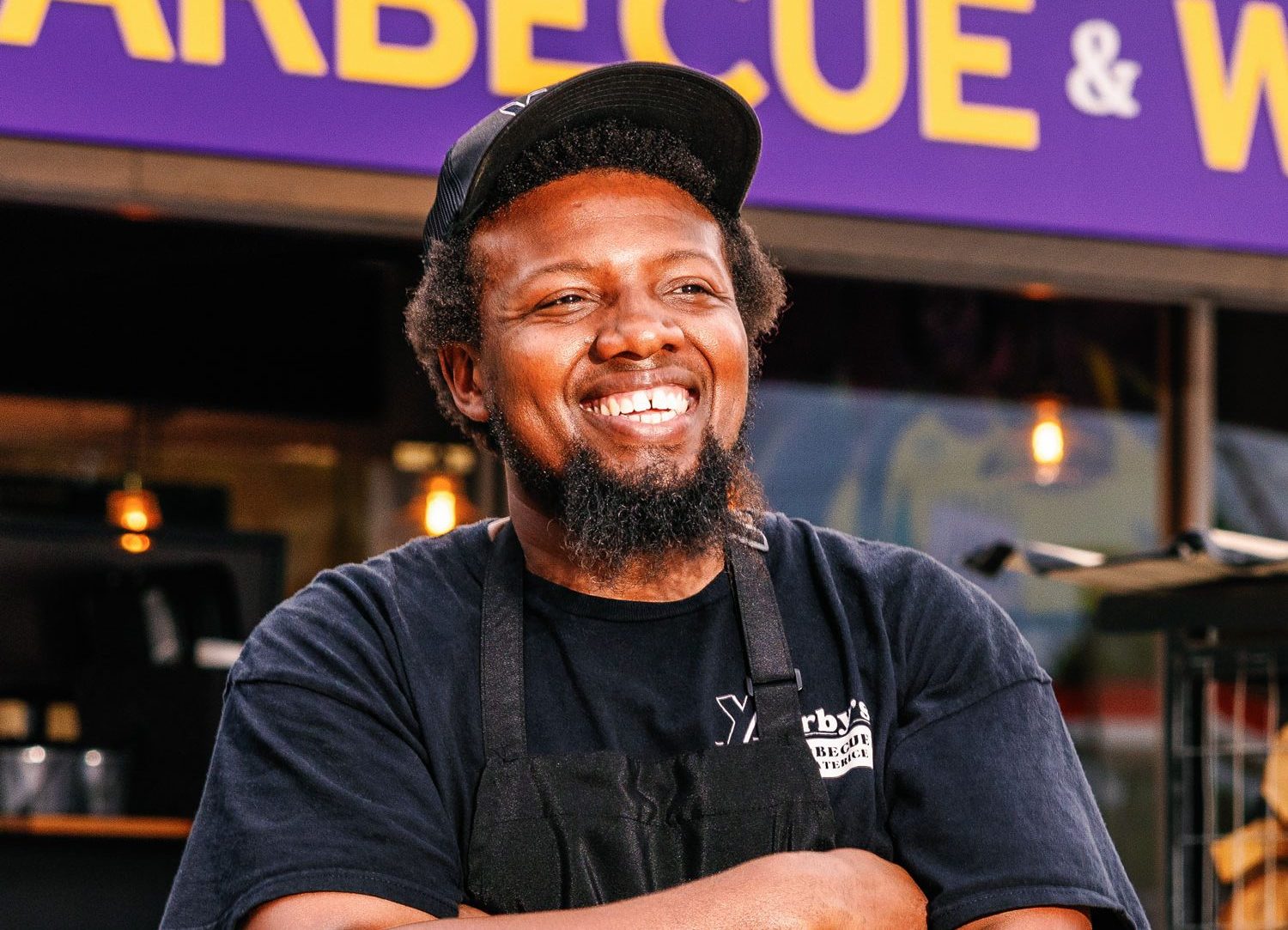 A Black man smiles with his arms crossed staring off to the side while wearing a black cap, shirt, and apron that reads "Yearby's"