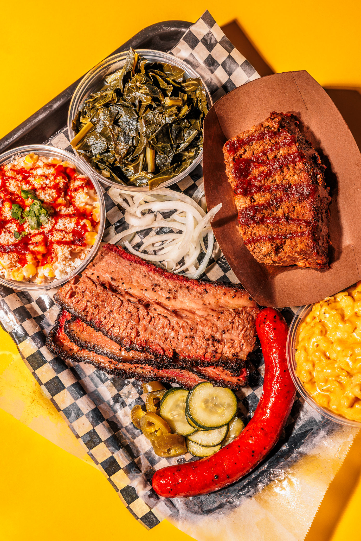A plate of assorted brisket, beef links, and meatloag along with macaroni and cheese, greens, and other sides on a checkered paper on a yellow table