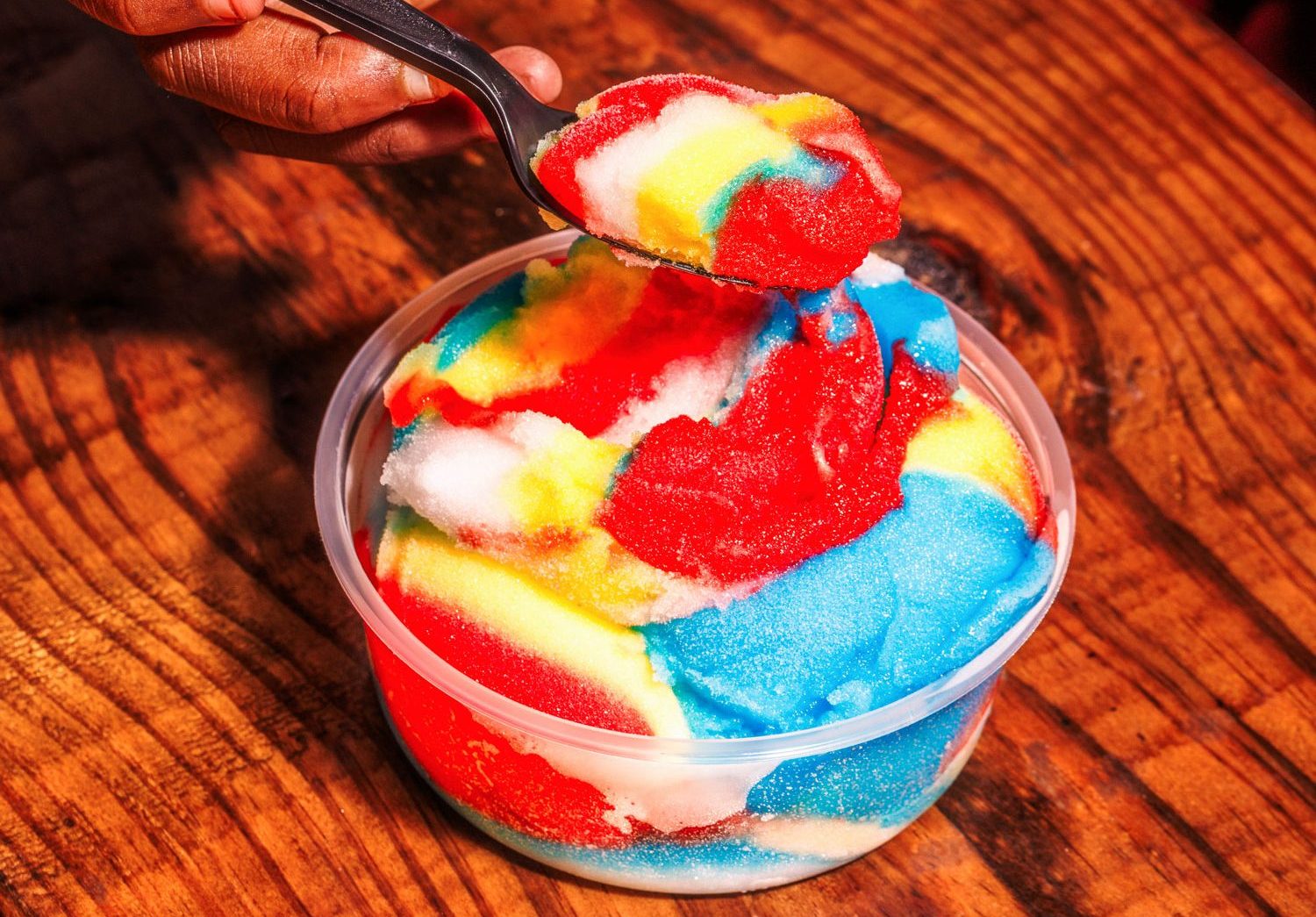 A hand holds a scoop of rainbow ice over a cup of rainbow ice