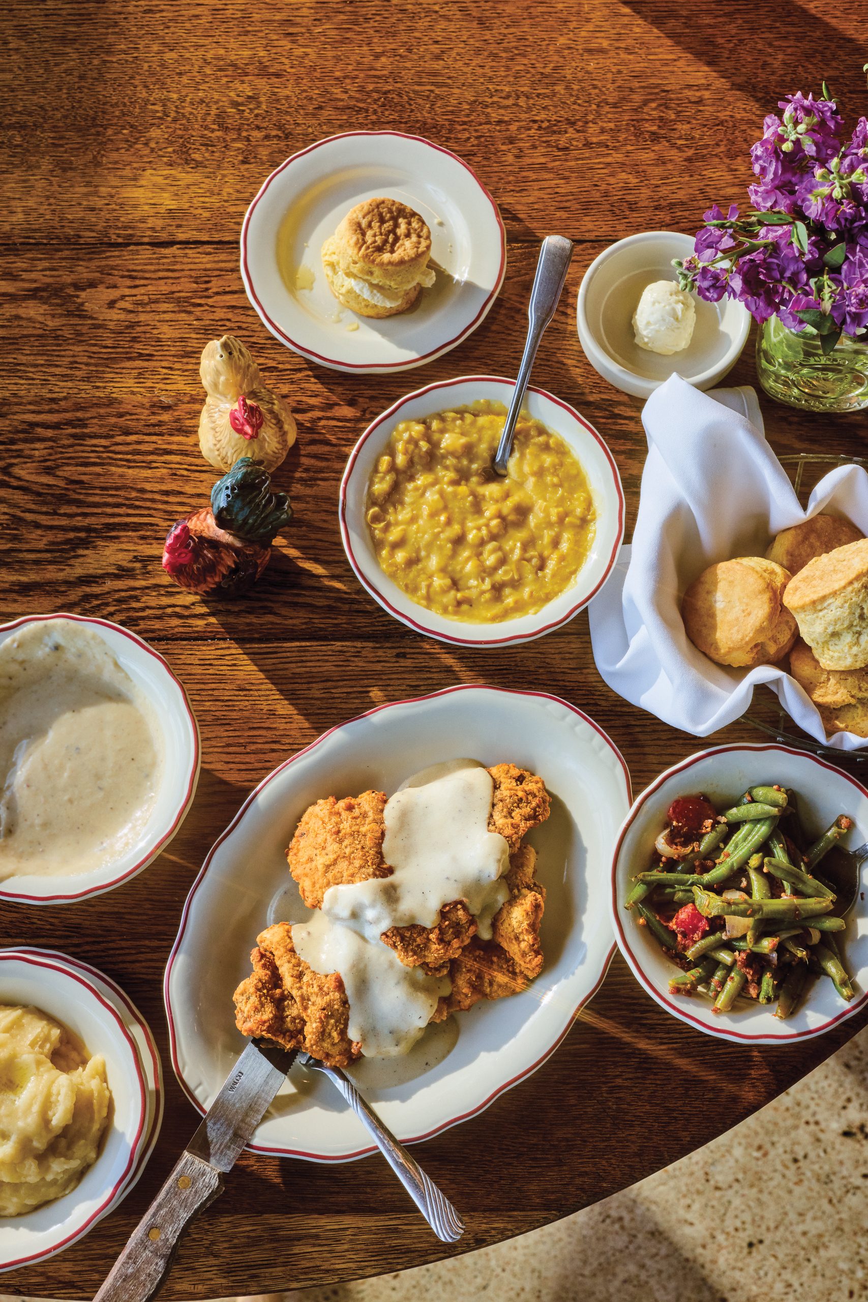 A spread of fried chicken, corn, biscuits, mashed potatoes, gravy, and green beans on a wood table, with a jar of lilacs placed nearby.