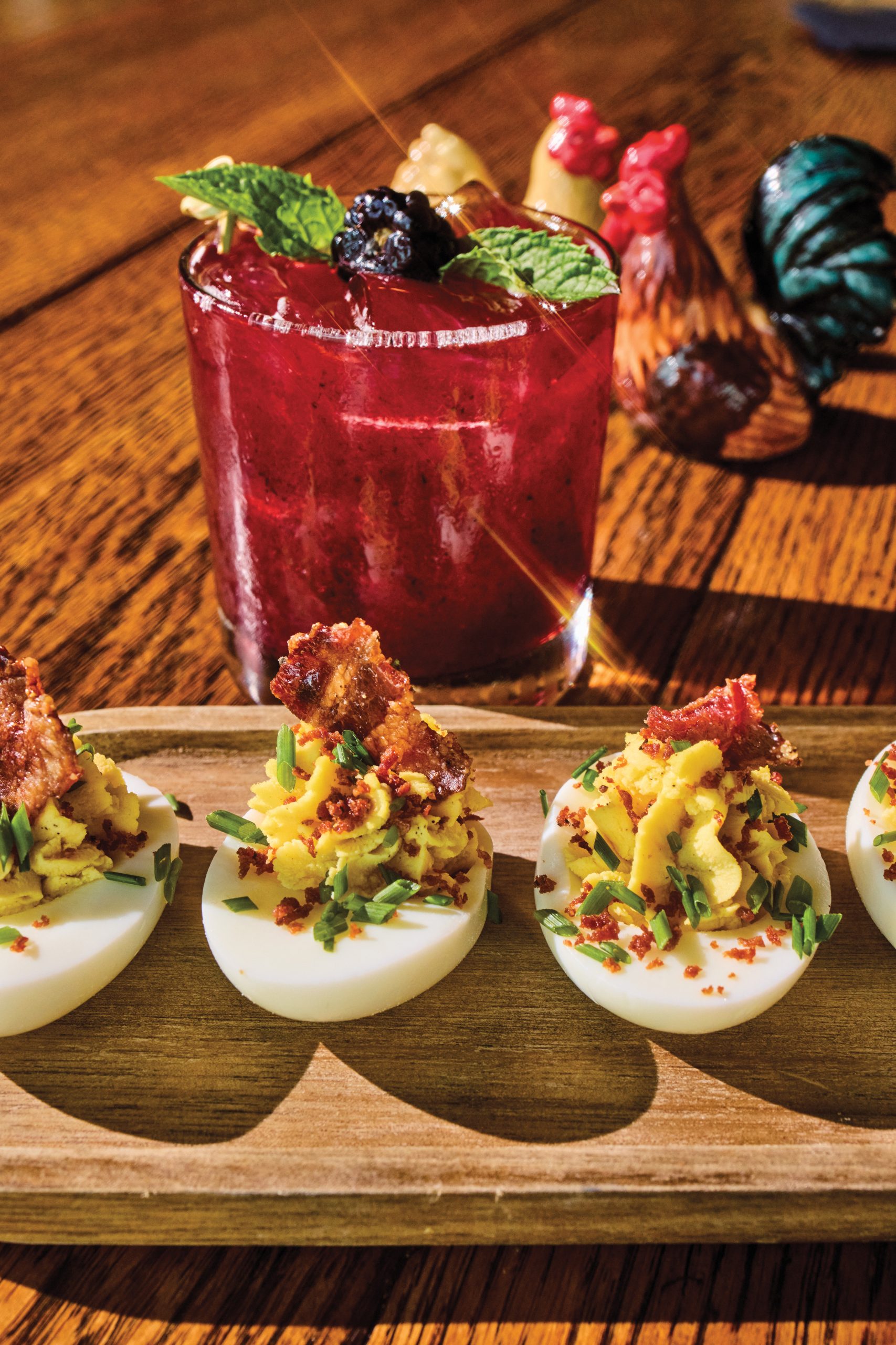 Four deviled eggs topped with chives and bacon on a wood plate in front of a berry cocktail. A pair of rooster and hen ceramic figures sits behind them.